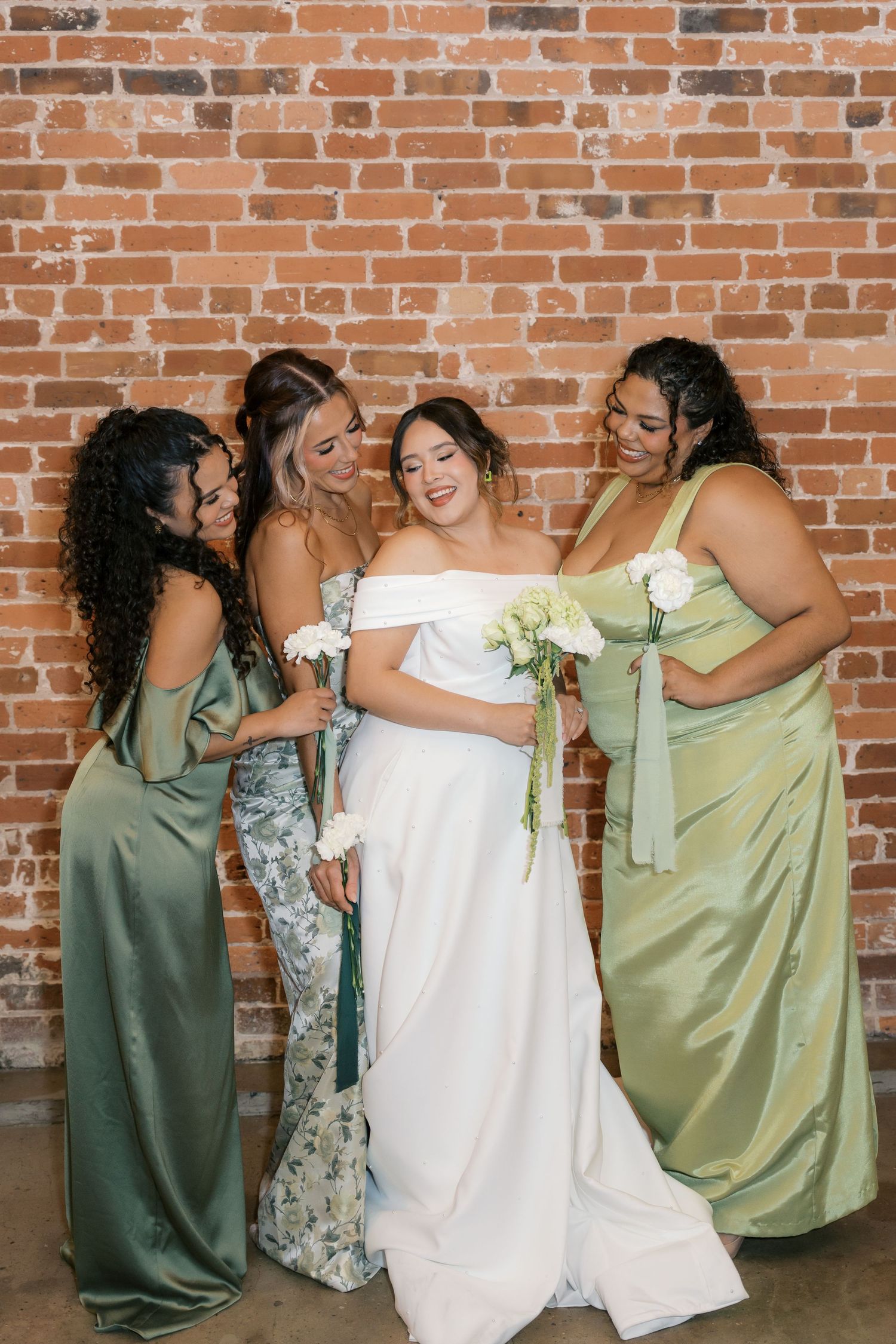 Wedding party in sage green and white attire shares candid moments against a brick wall backdrop.