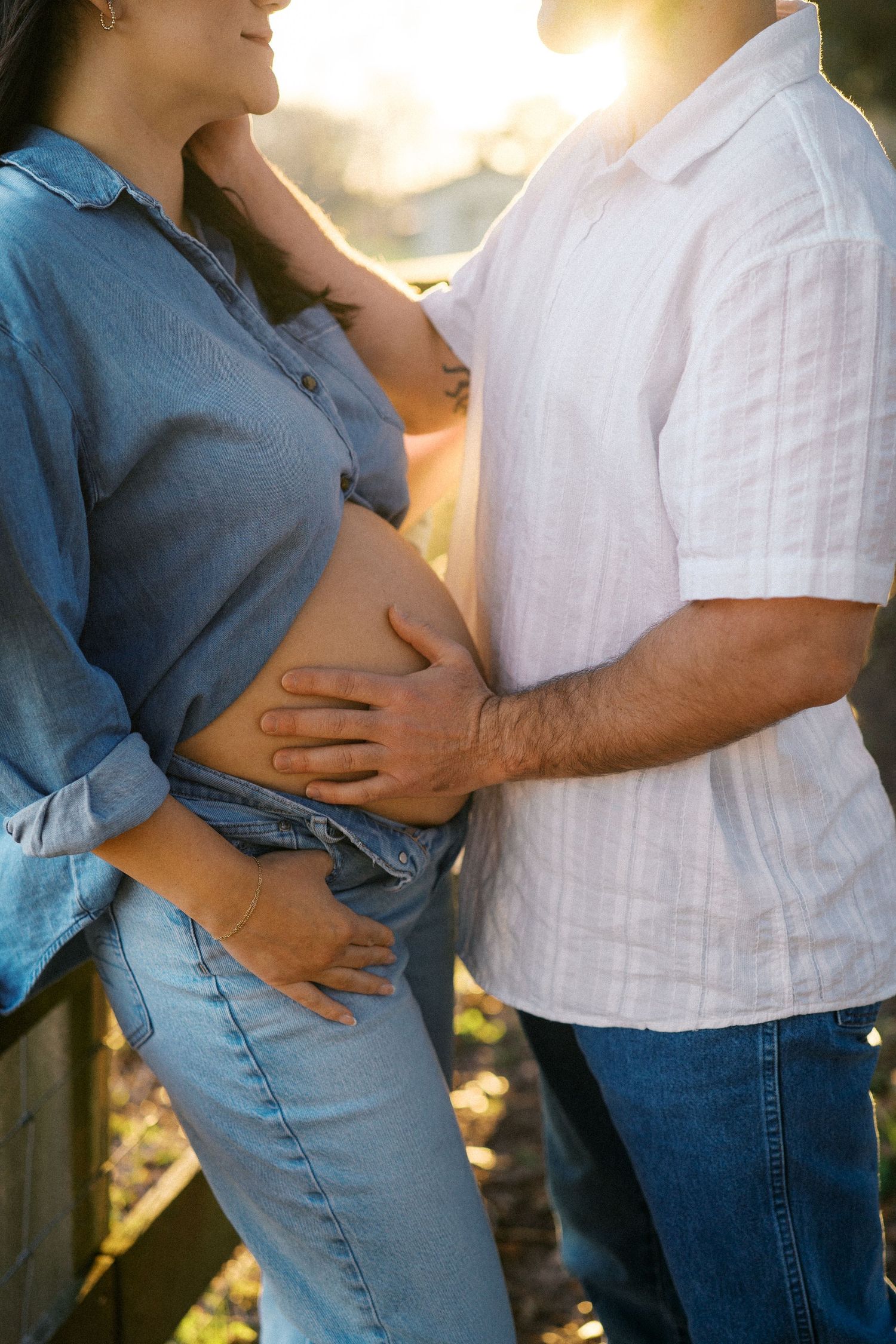 A couple in casual clothing stands together in sunlight as they lovingly touch a pregnant belly.