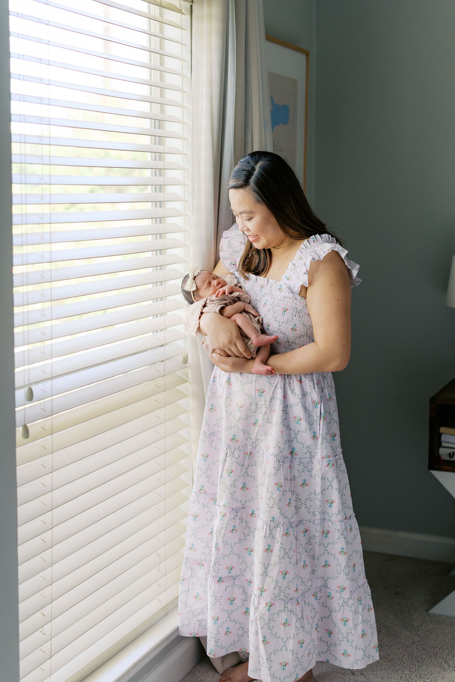 Mother in floral dress cradles newborn baby near window with natural light.