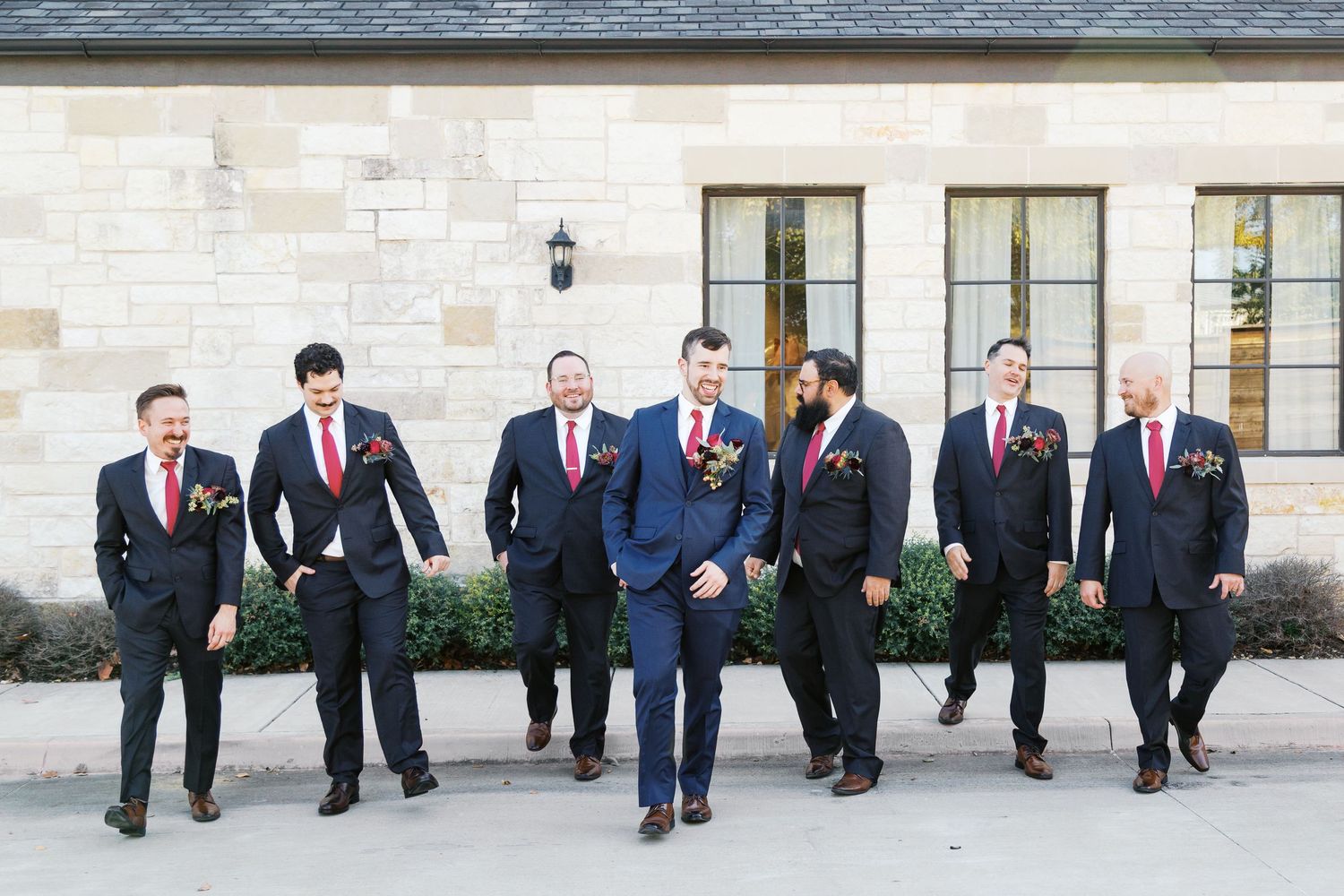 Groomsmen in dark suits posing casually outside stone venue.