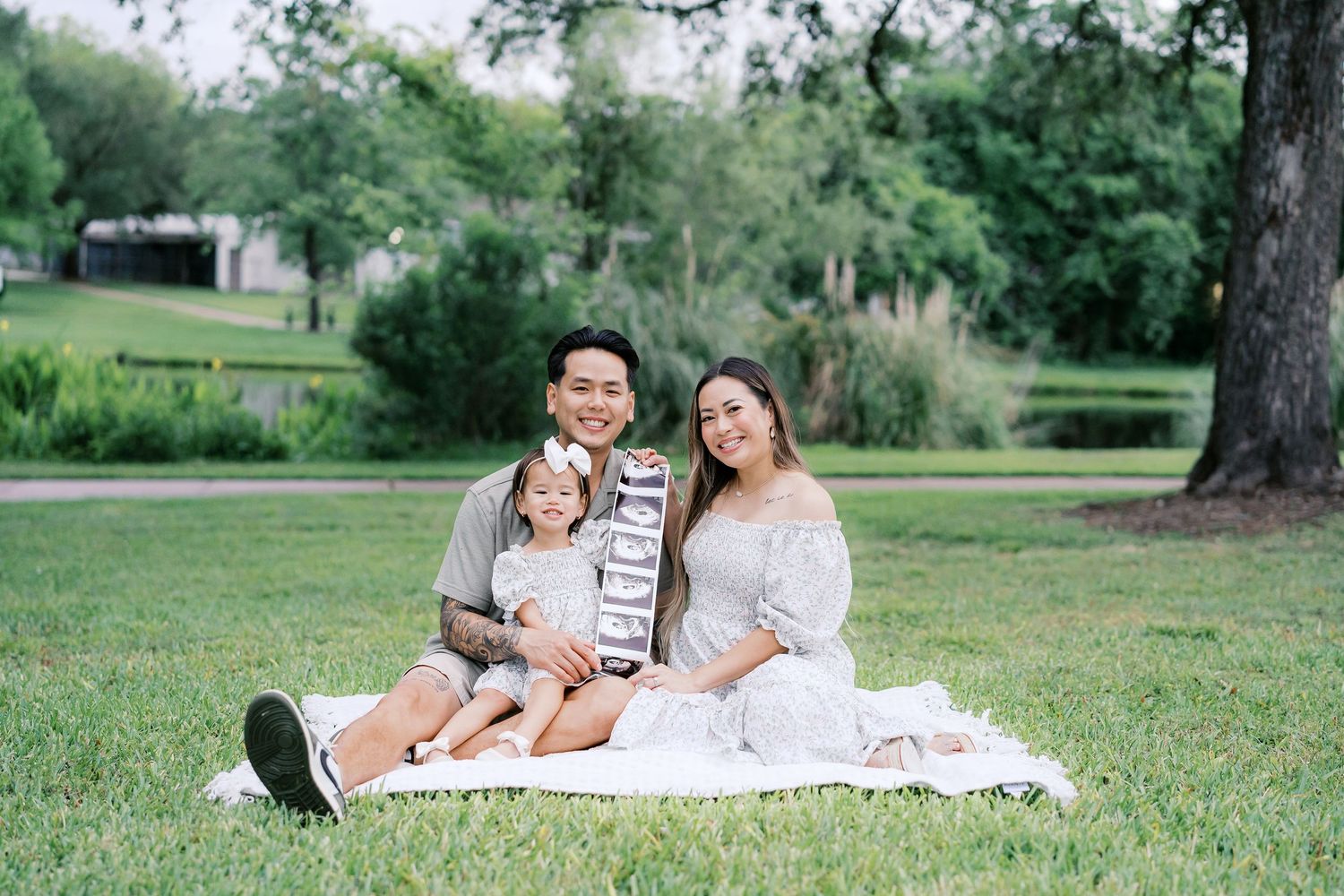 Happy Asian family of three sitting on a blanket in a lush green park, parents smiling with young child between them.