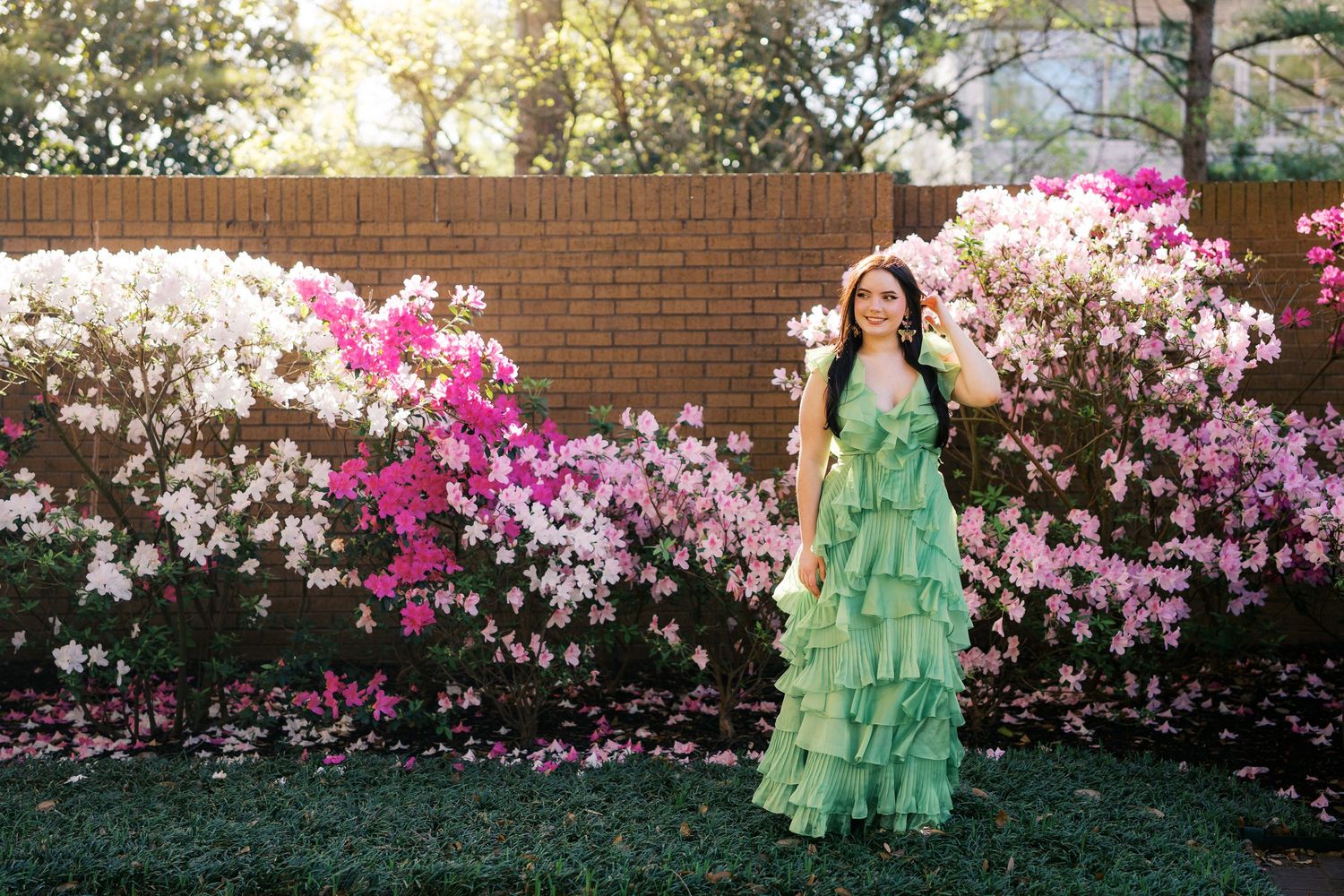 Woman in green ruffled dress poses in garden with blooming pink azalea bushes during spring photoshoot.