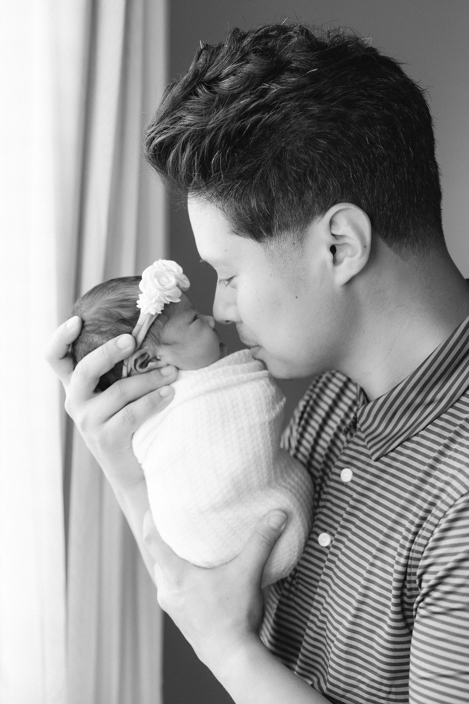 Black and white portrait of parent tenderly holding swaddled newborn with rose headband.