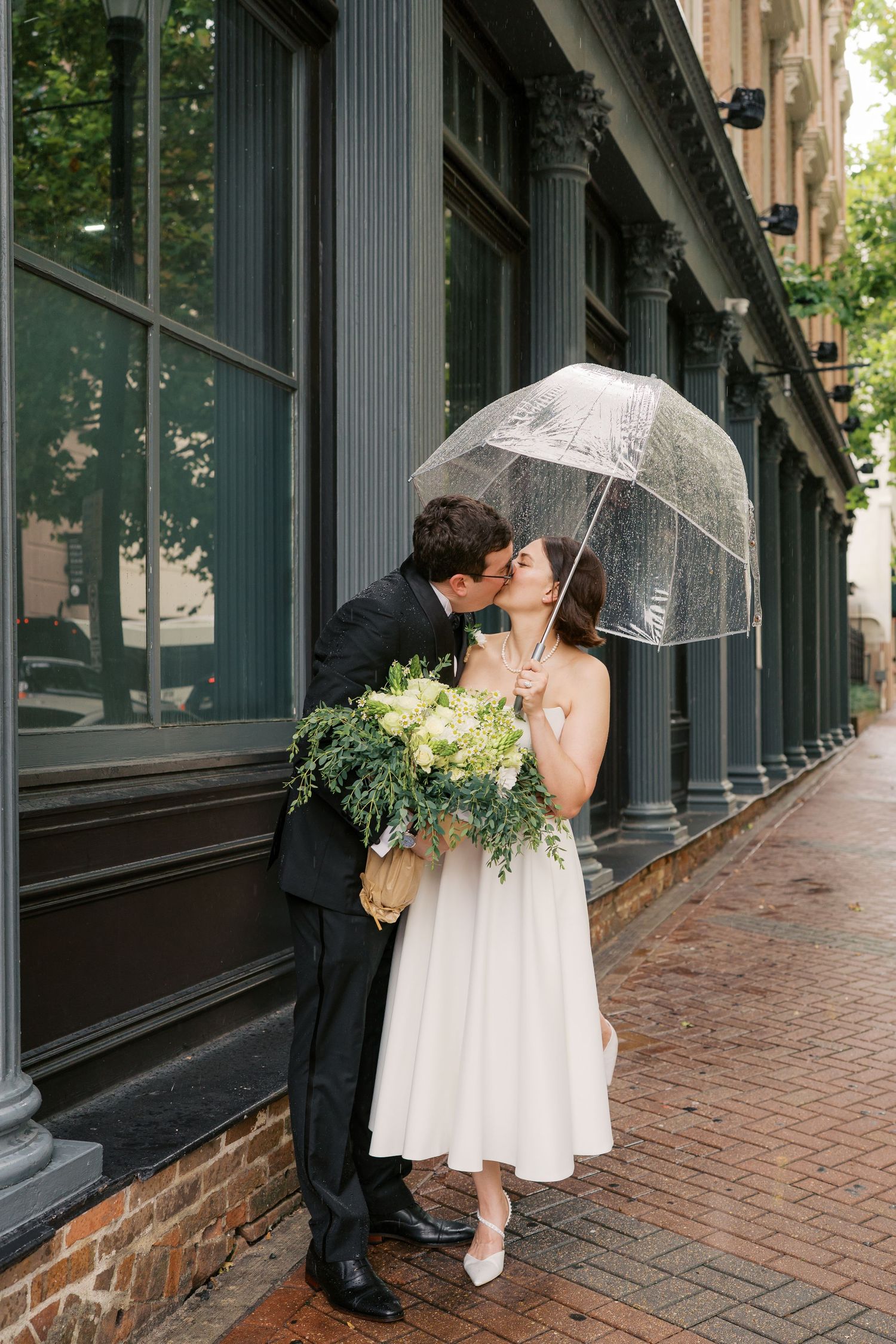 Romantic couple sharing a moment under clear umbrellas along a brick building.