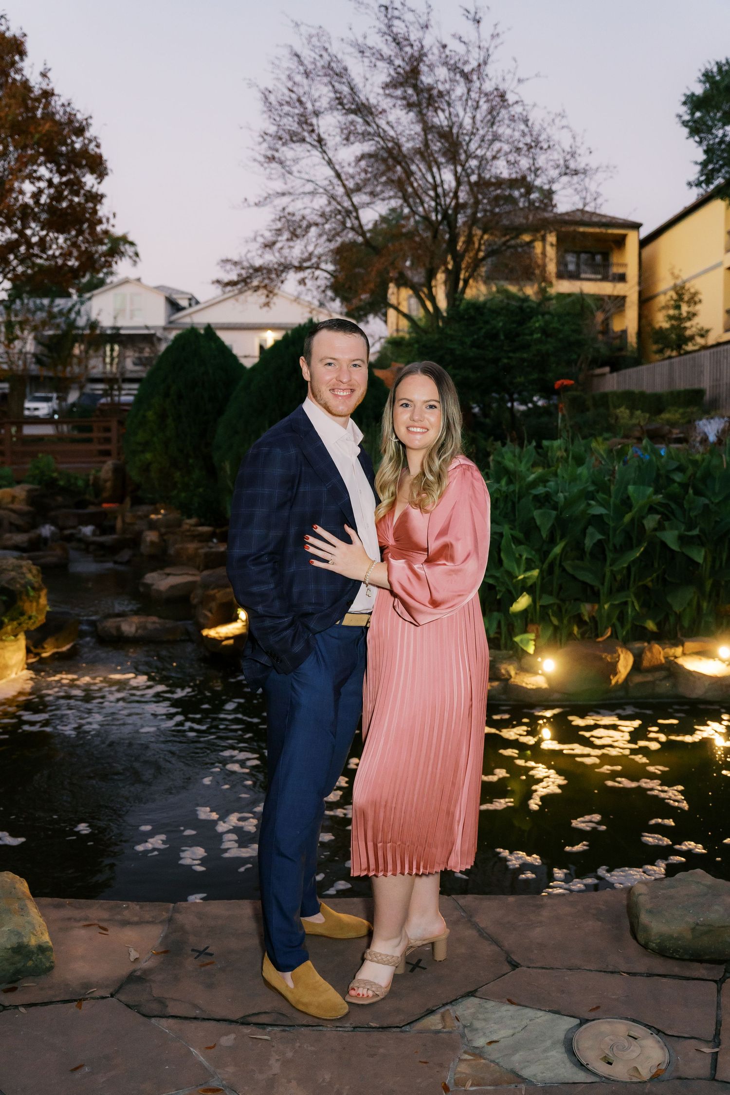 Couple poses by an illuminated pond at dusk for romantic engagement photos.