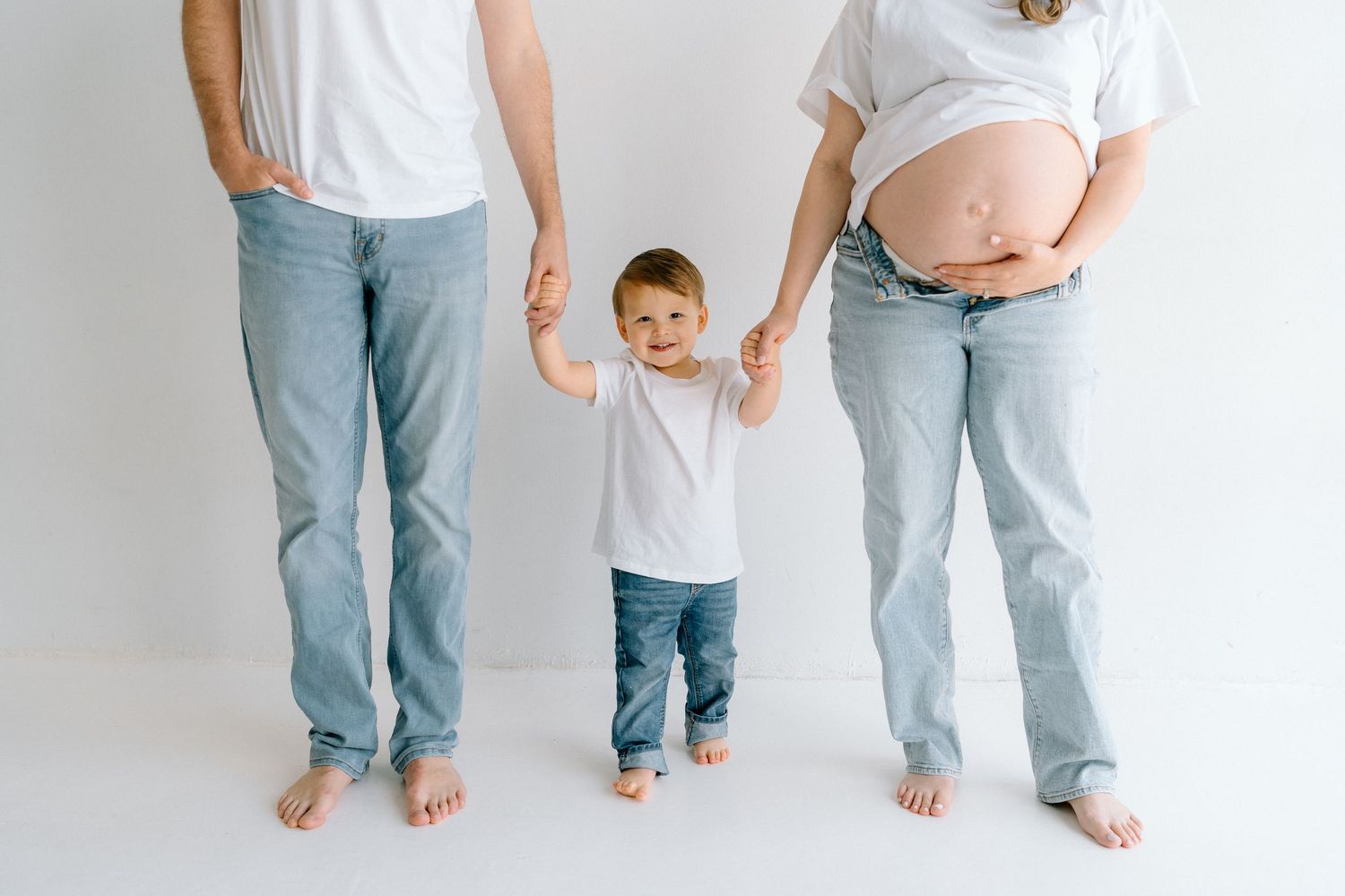 Casual family maternity session with coordinated outfits showing feet and legs against white background.