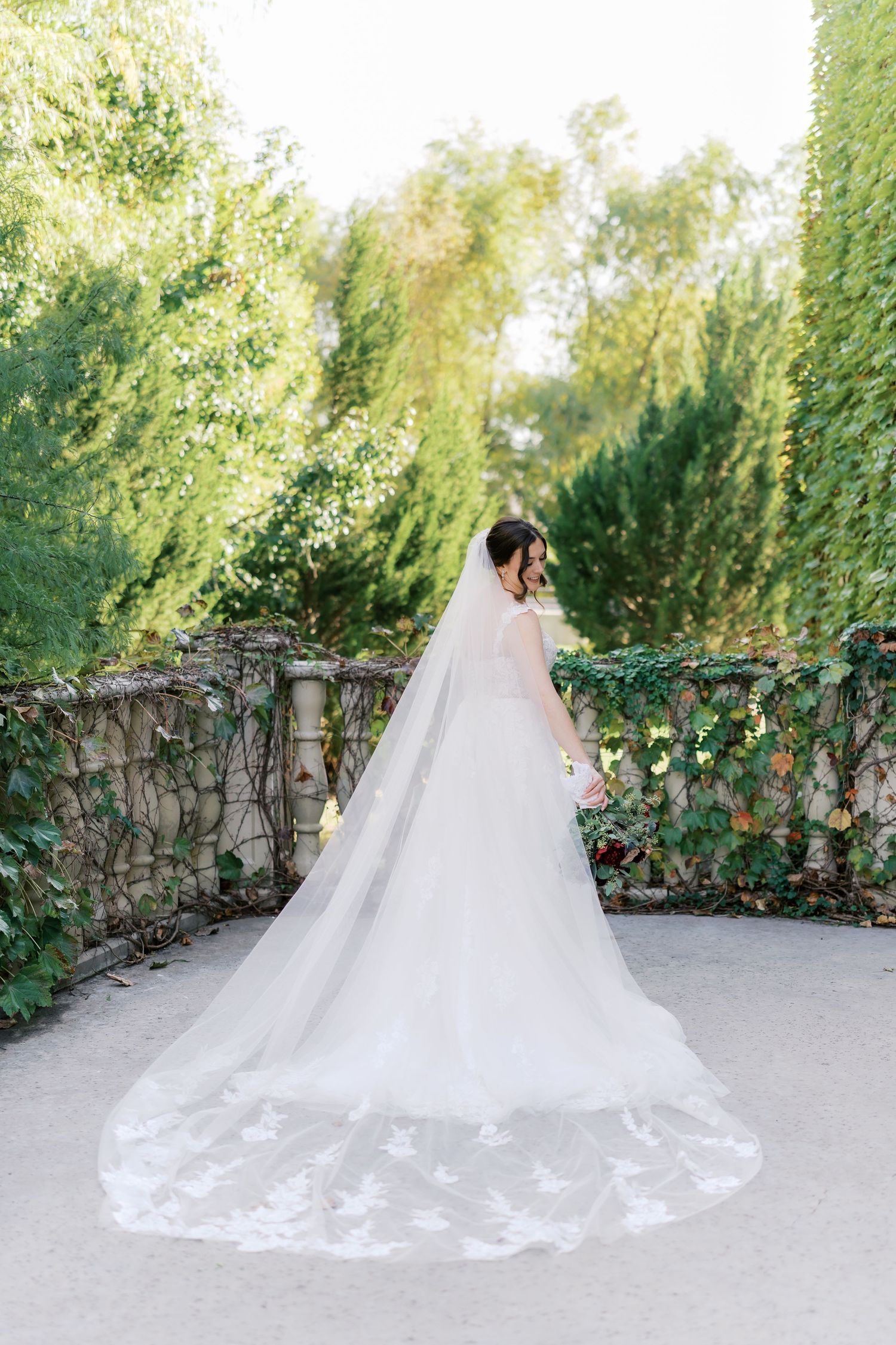 Wedding dress with long lace train displayed in garden setting with vine-covered wall.