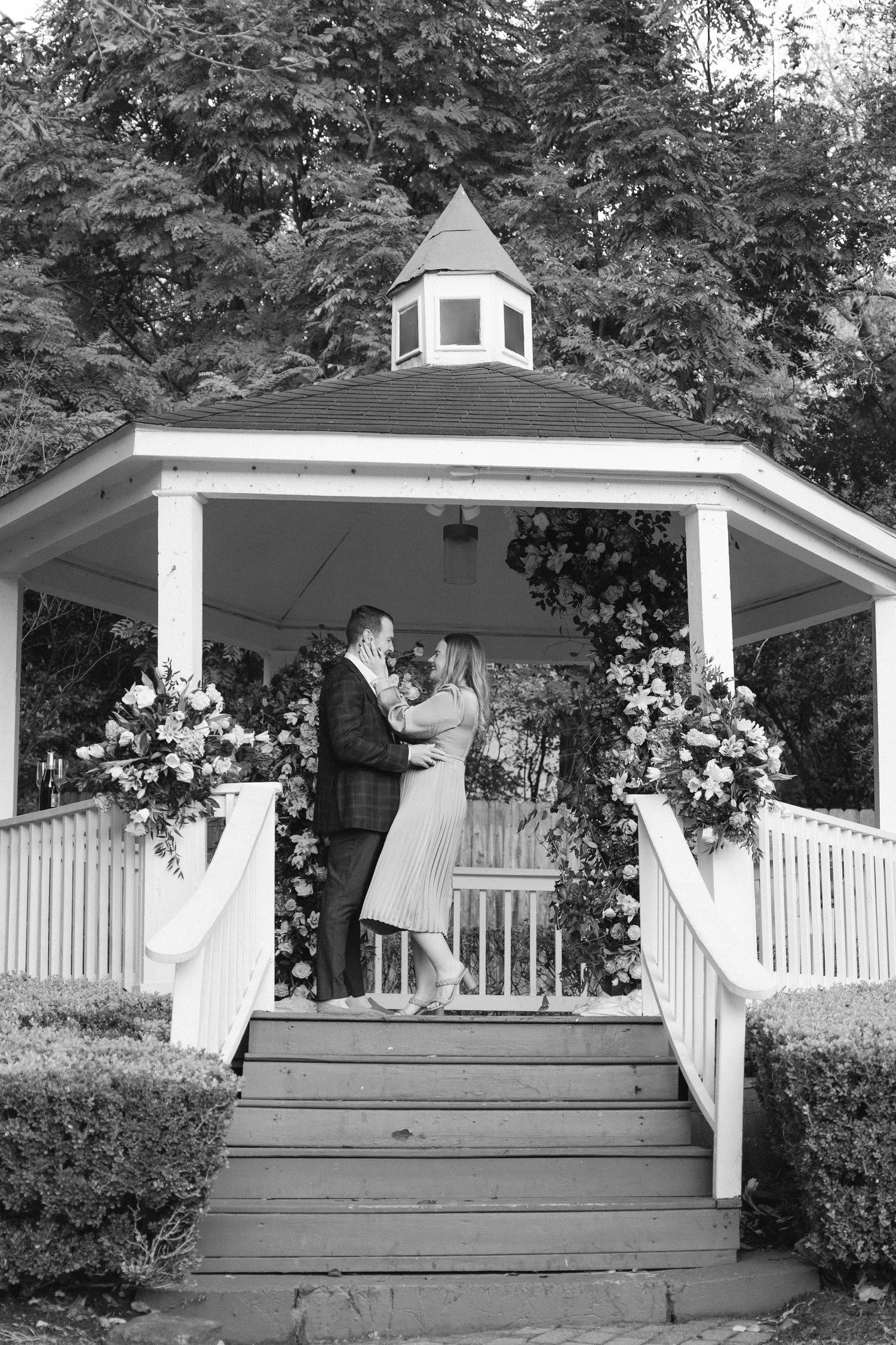 A black and white capture of a romantic proposal scene on a decorated garden gazebo.