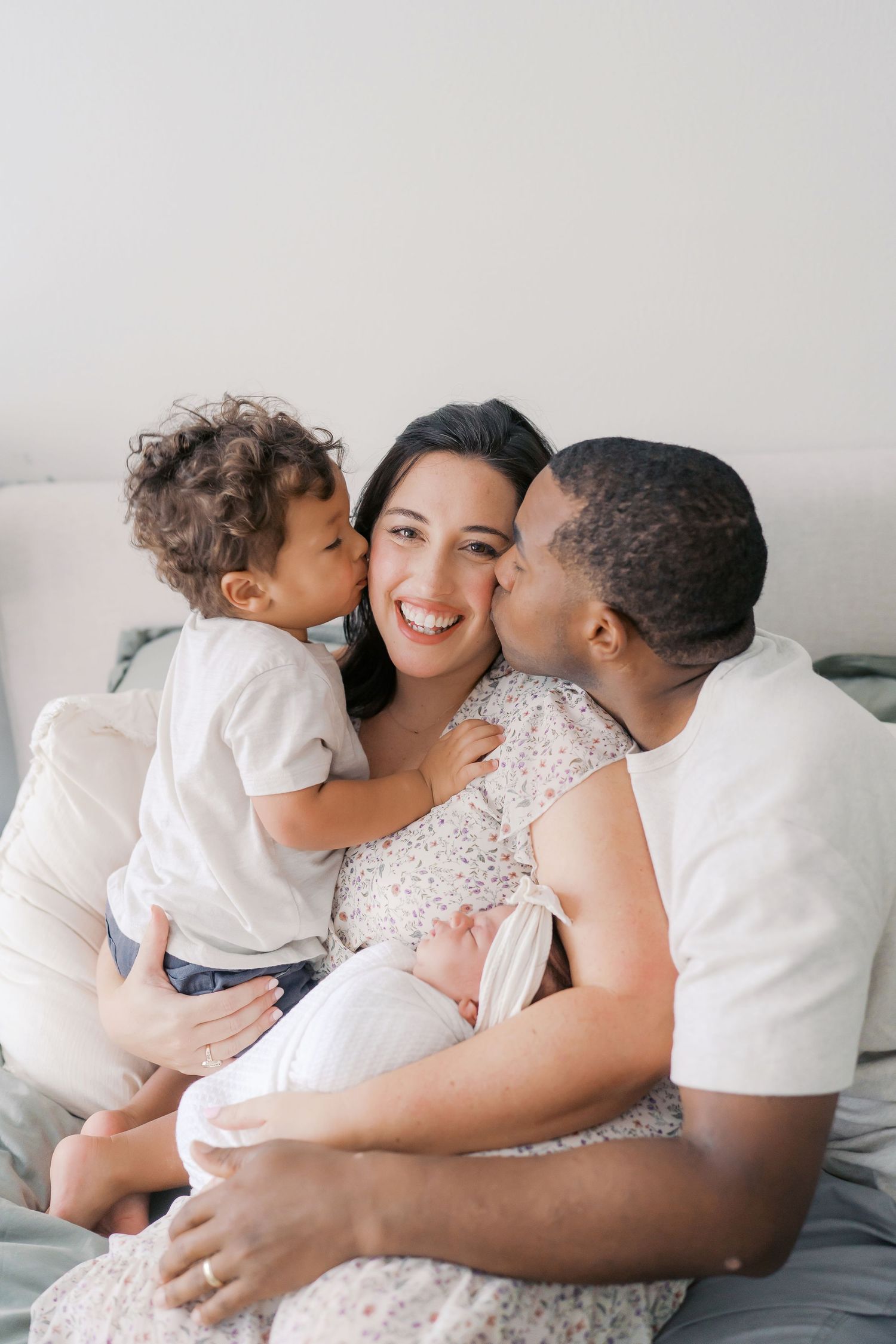 A happy family with a toddler and newborn sharing kisses on a bed in a bright, cozy room.