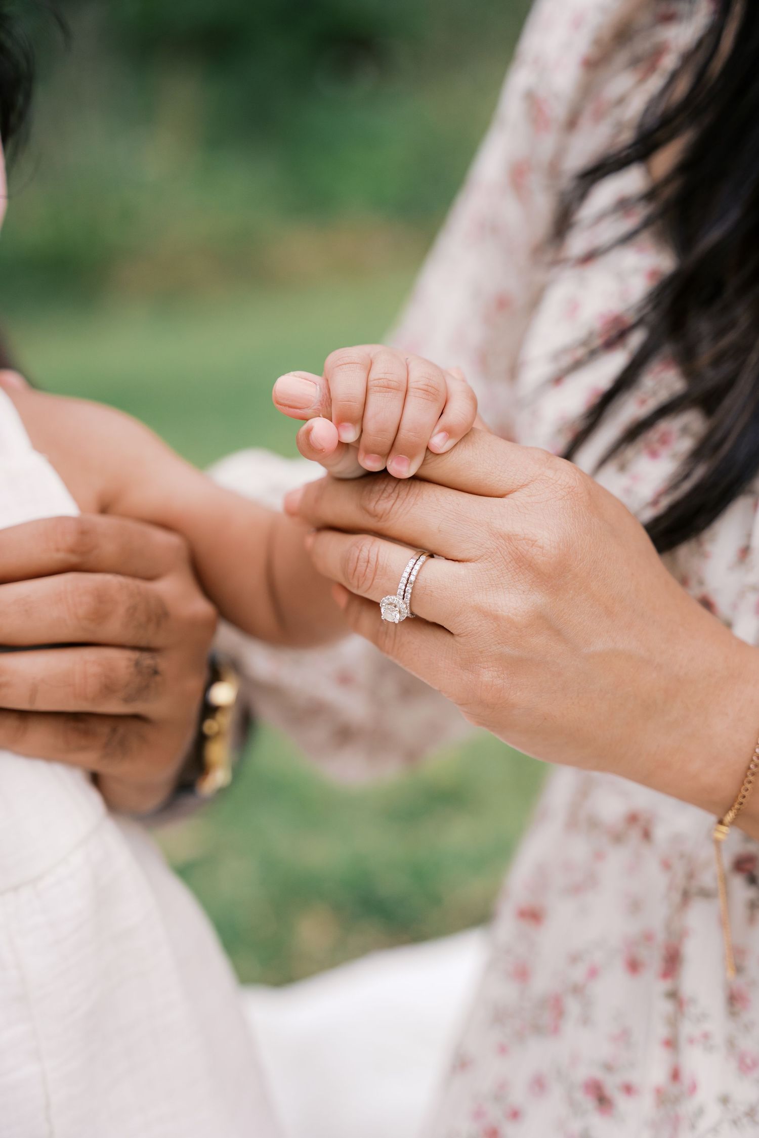 Close-up of woman's hand wearing diamond engagement ring while holding a baby's tiny hand outdoors.