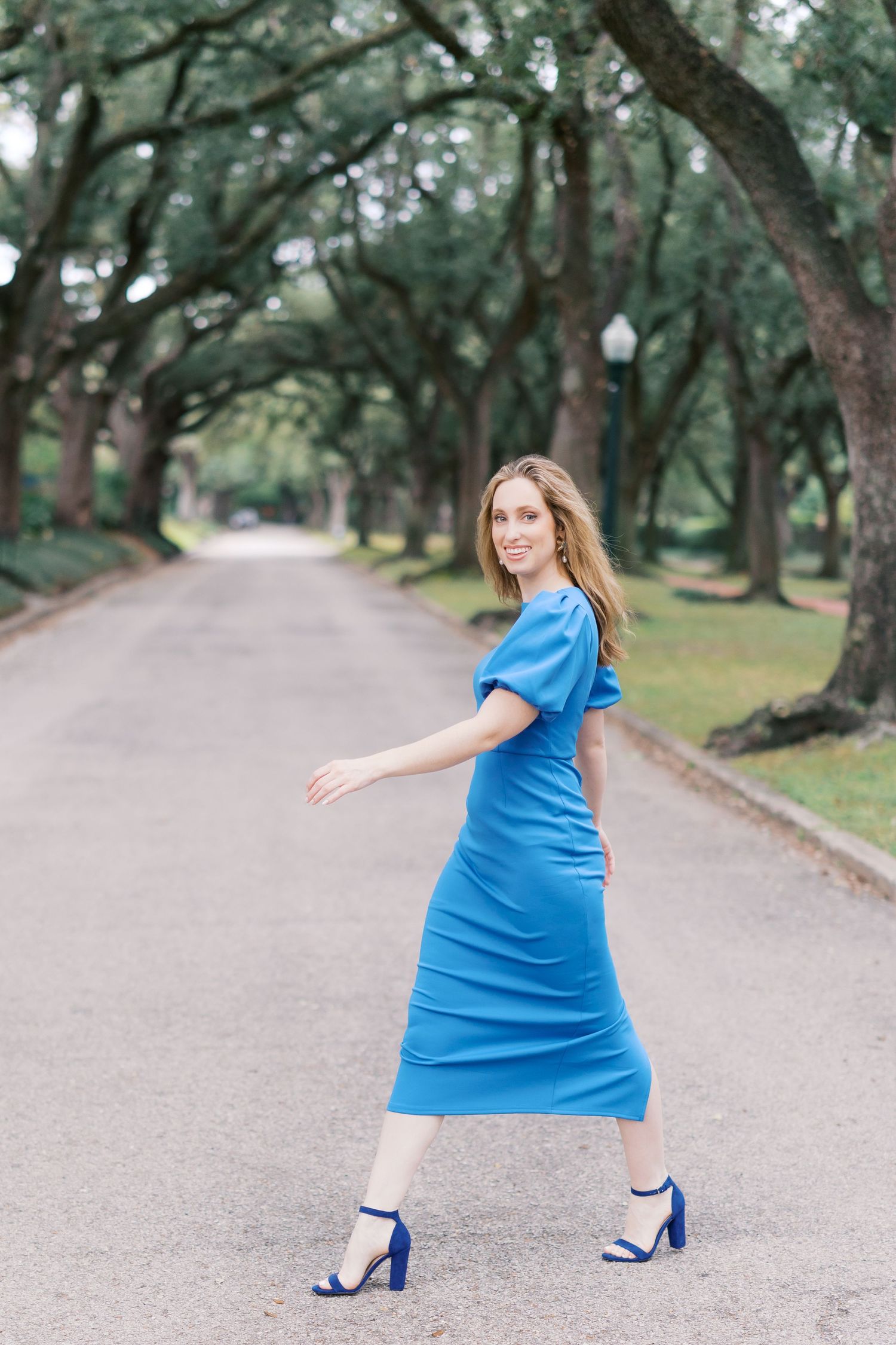 A person wearing a vibrant blue dress poses gracefully on a gravel walkway surrounded by trees.