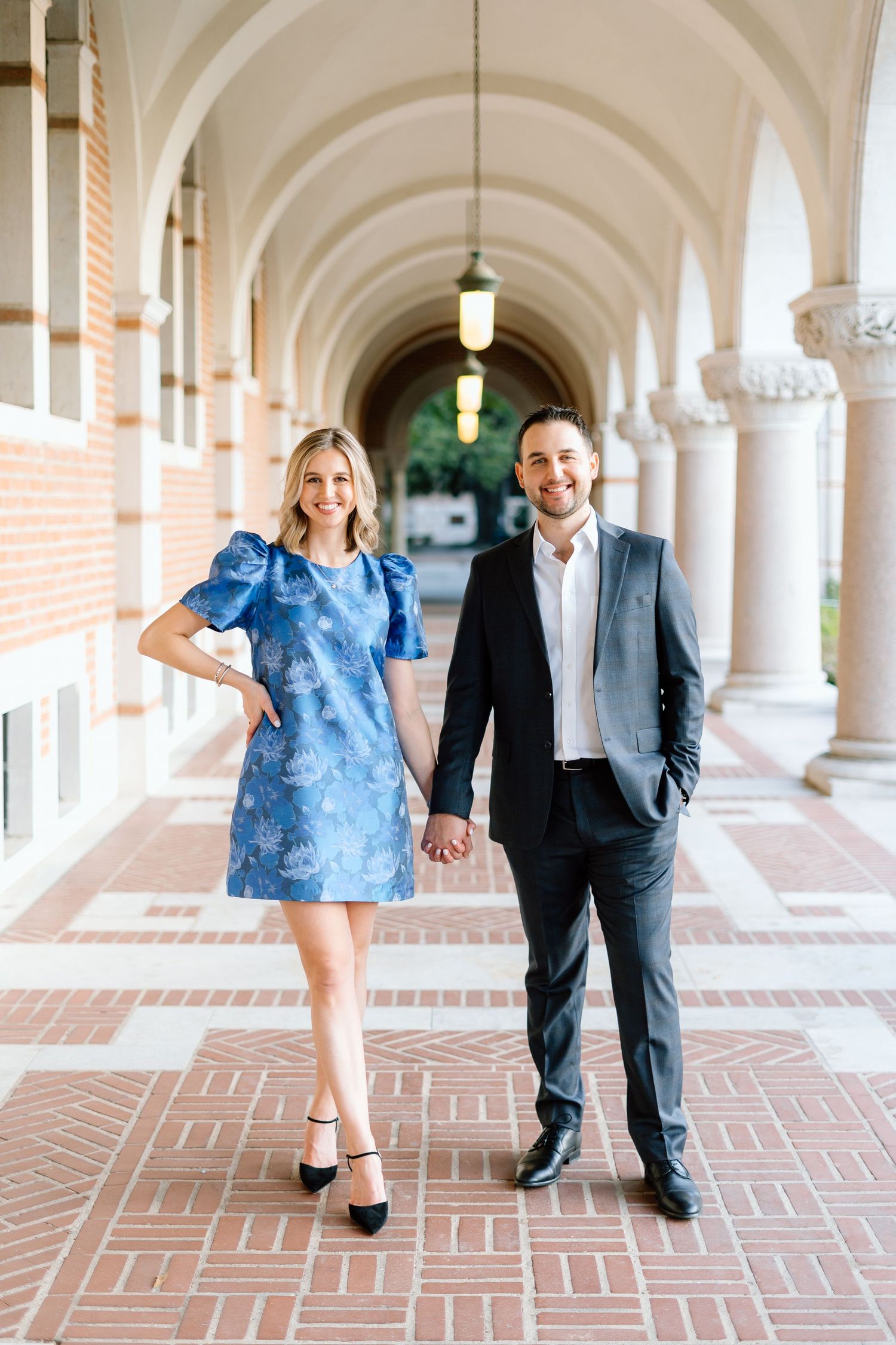 A couple walks and poses playfully along a covered walkway with arched columns.