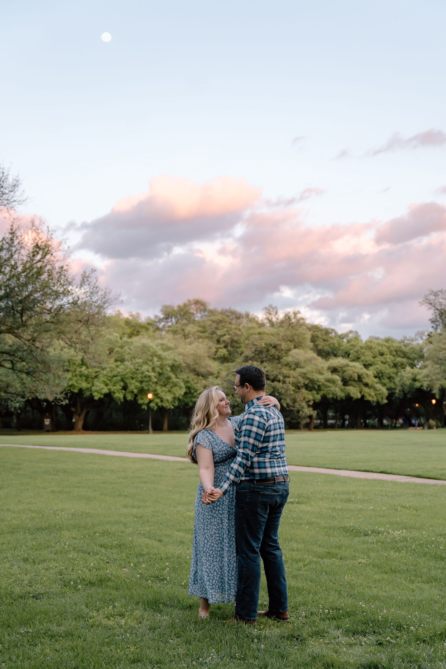 Couple standing close together in a grassy field during sunset with trees in the background.