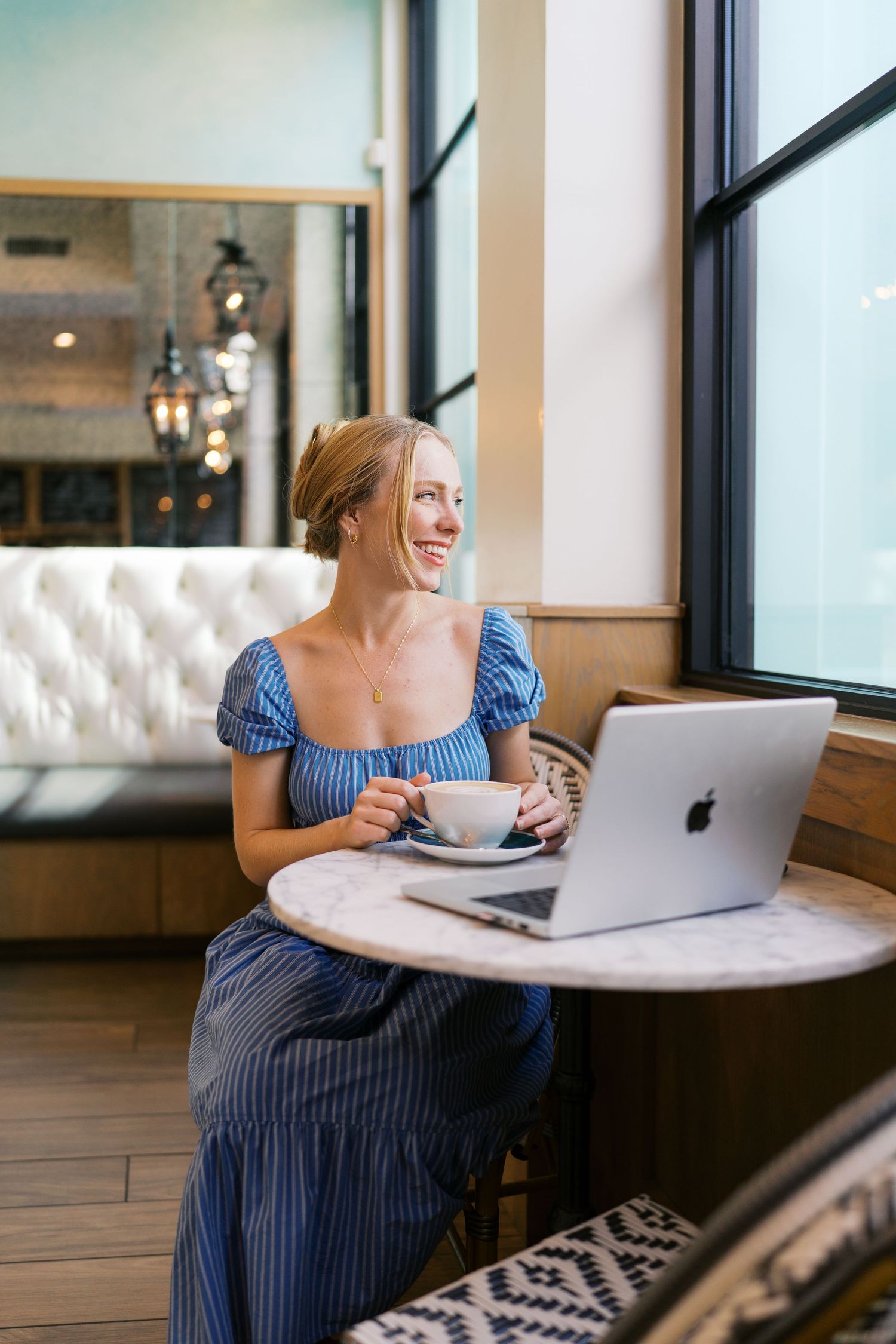 Woman in blue dress works on laptop by window in modern cafe setting.