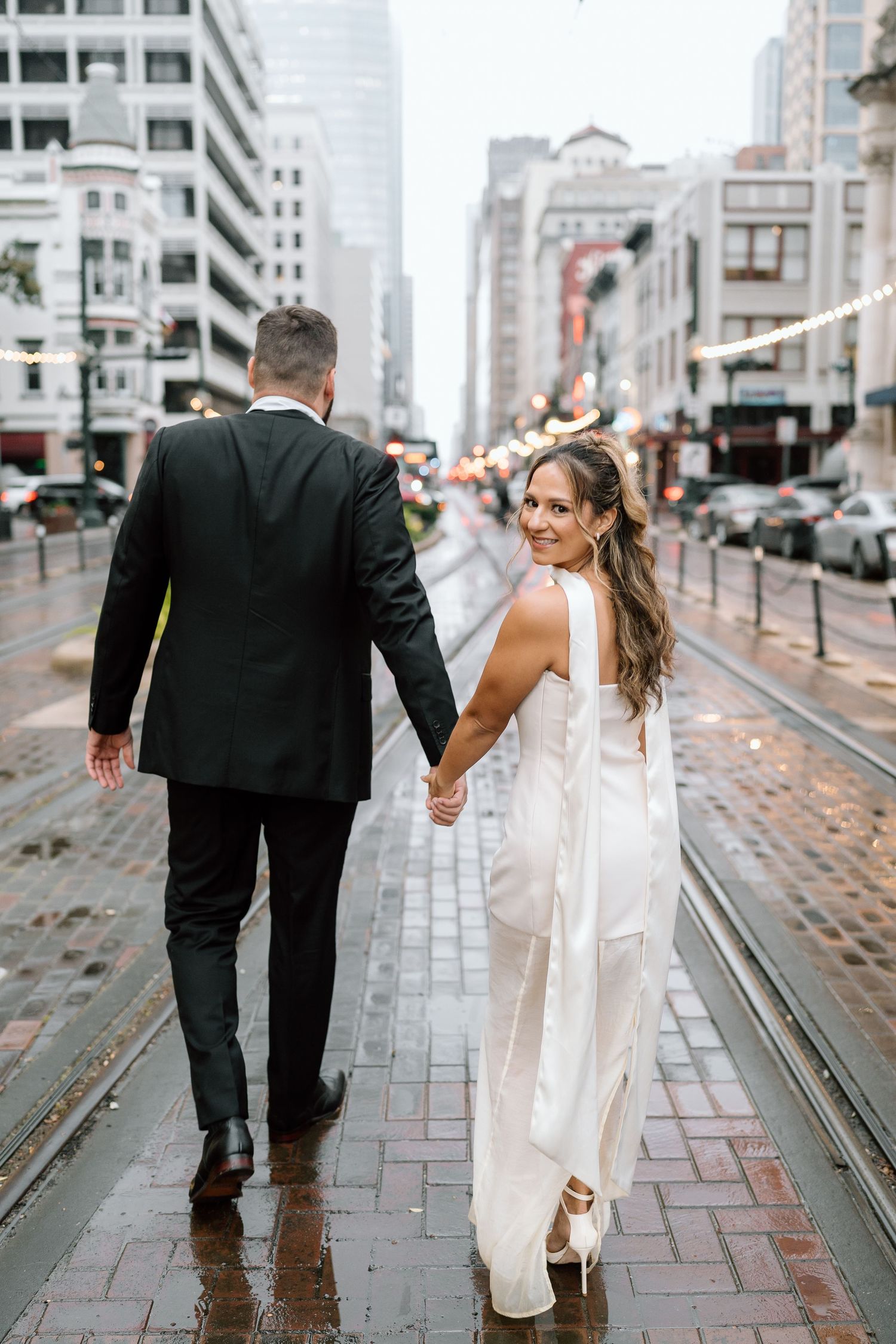Couple walks hand in hand along urban trolley tracks on rainy day.