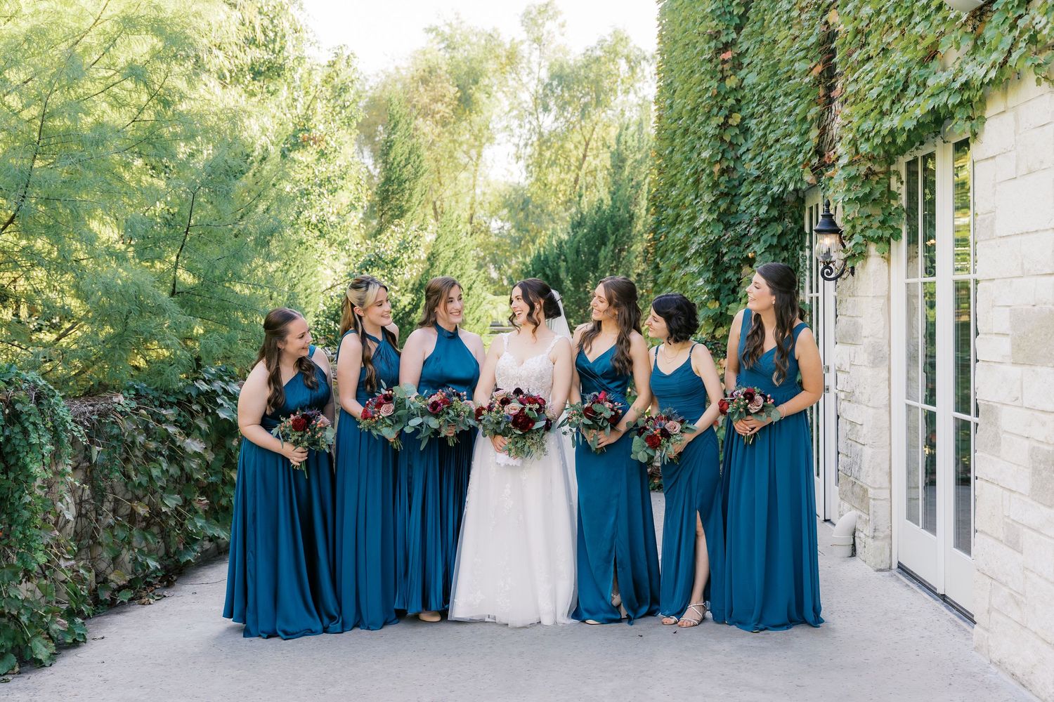 Bridal party in teal blue dresses pose together against stone and ivy wall.
