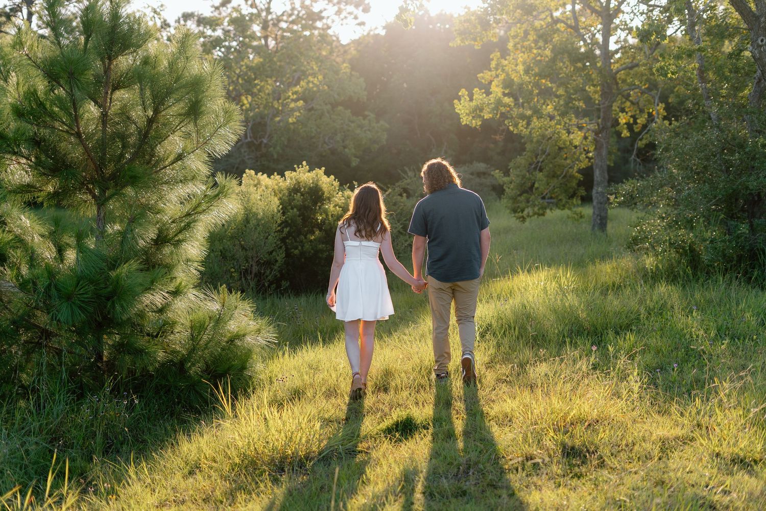 Couple walking through a sunlit meadow at golden hour with trees in the background.
