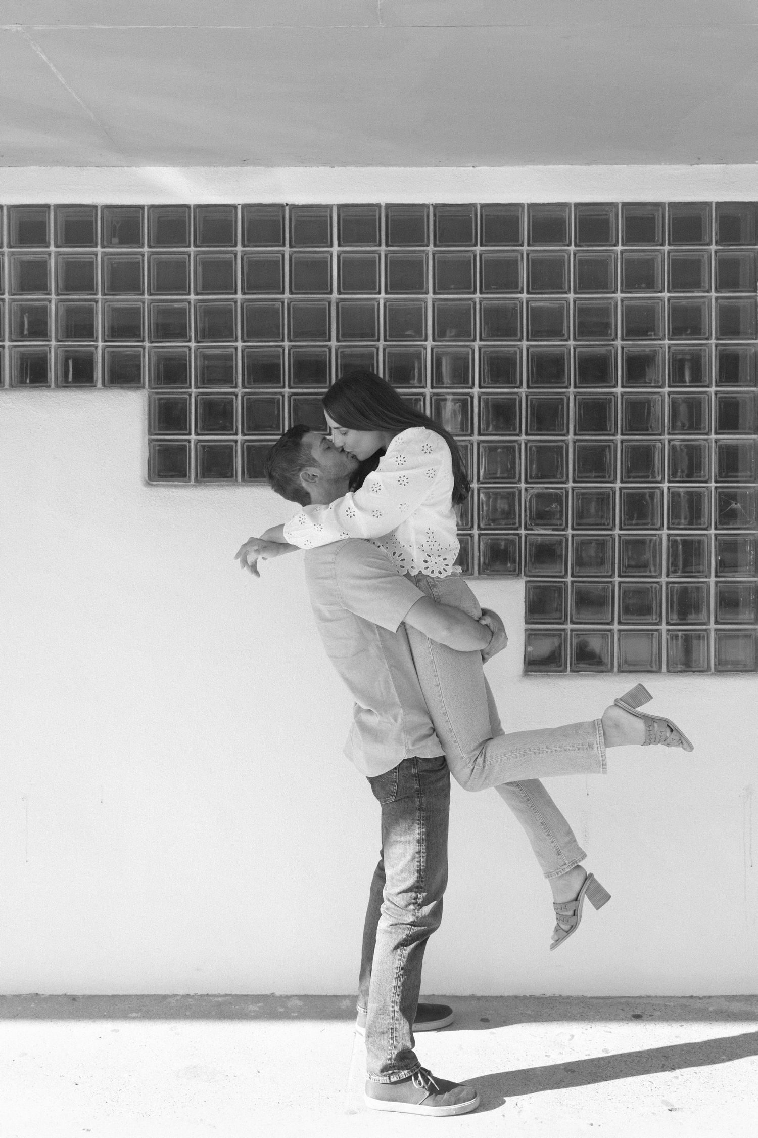 A couple poses playfully against a white wall with glass block windows.