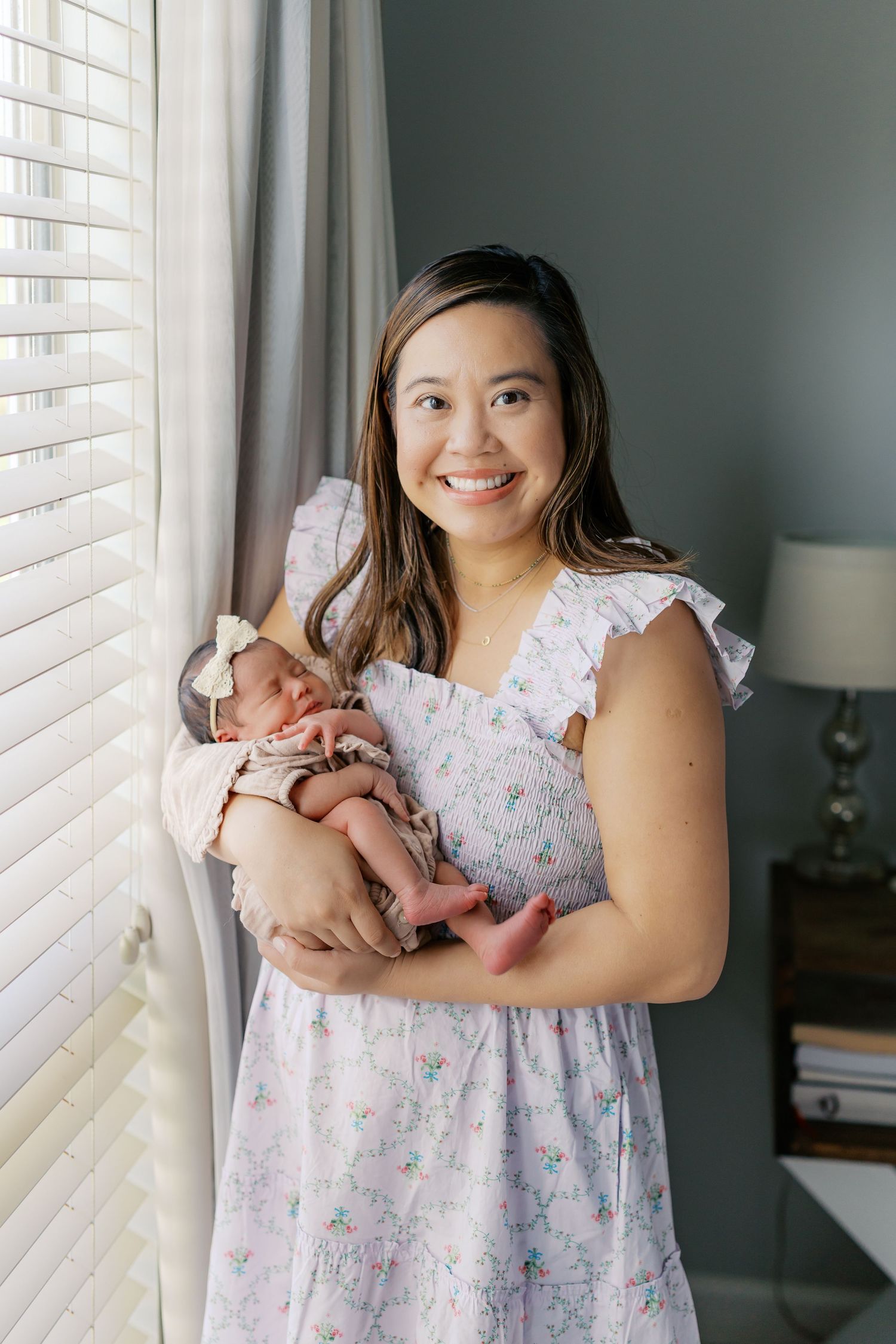Woman in floral dress holds newborn baby by window with white blinds.