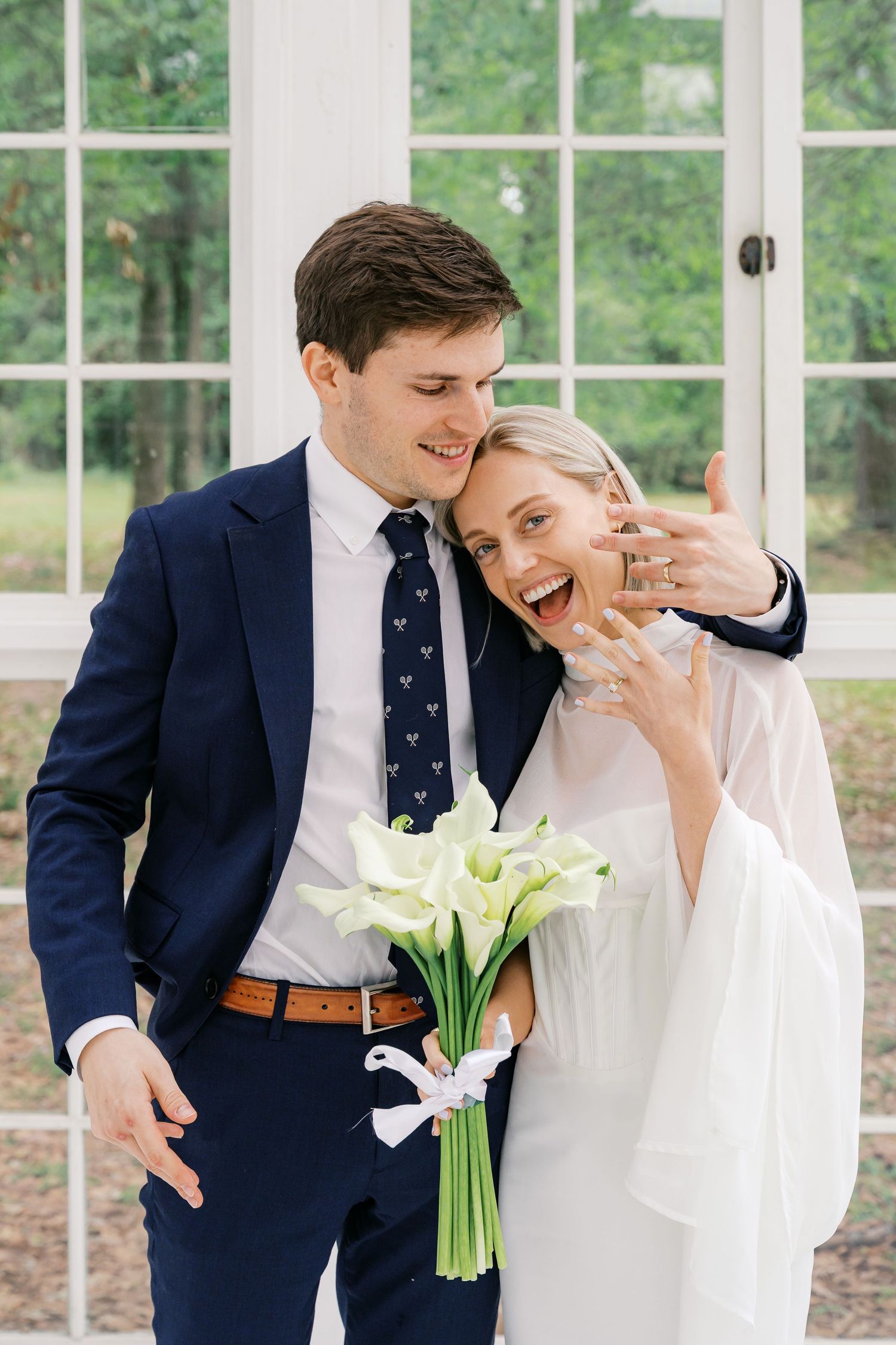 Joyful bride shows off wedding ring while groom holds calla lily bouquet, posing together in white wedding attire.