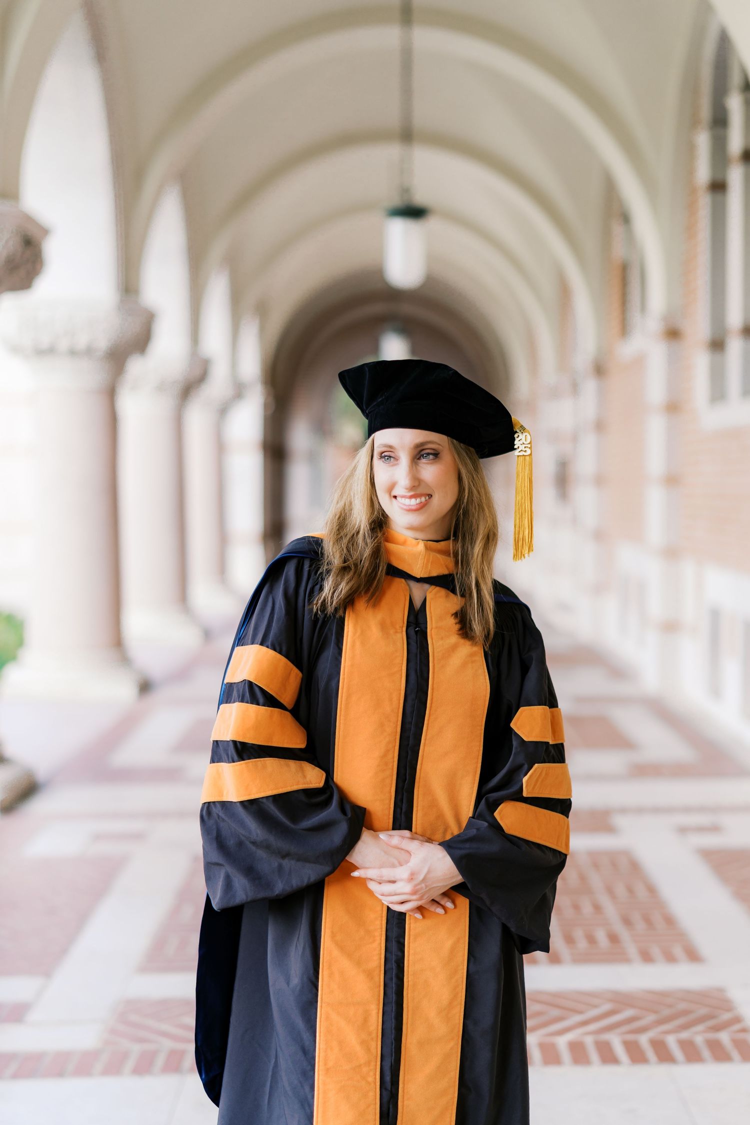 Graduate in doctoral regalia with orange trim smiles in historic university hallway.