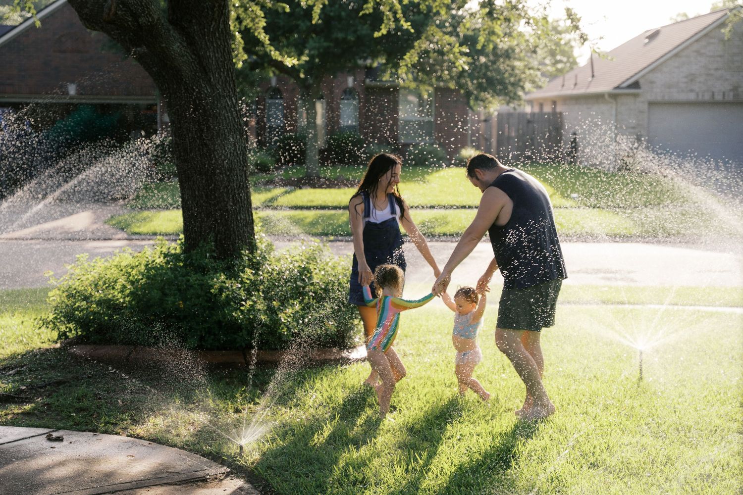 Family playing in sprinklers on green lawn beside large tree on sunny day.