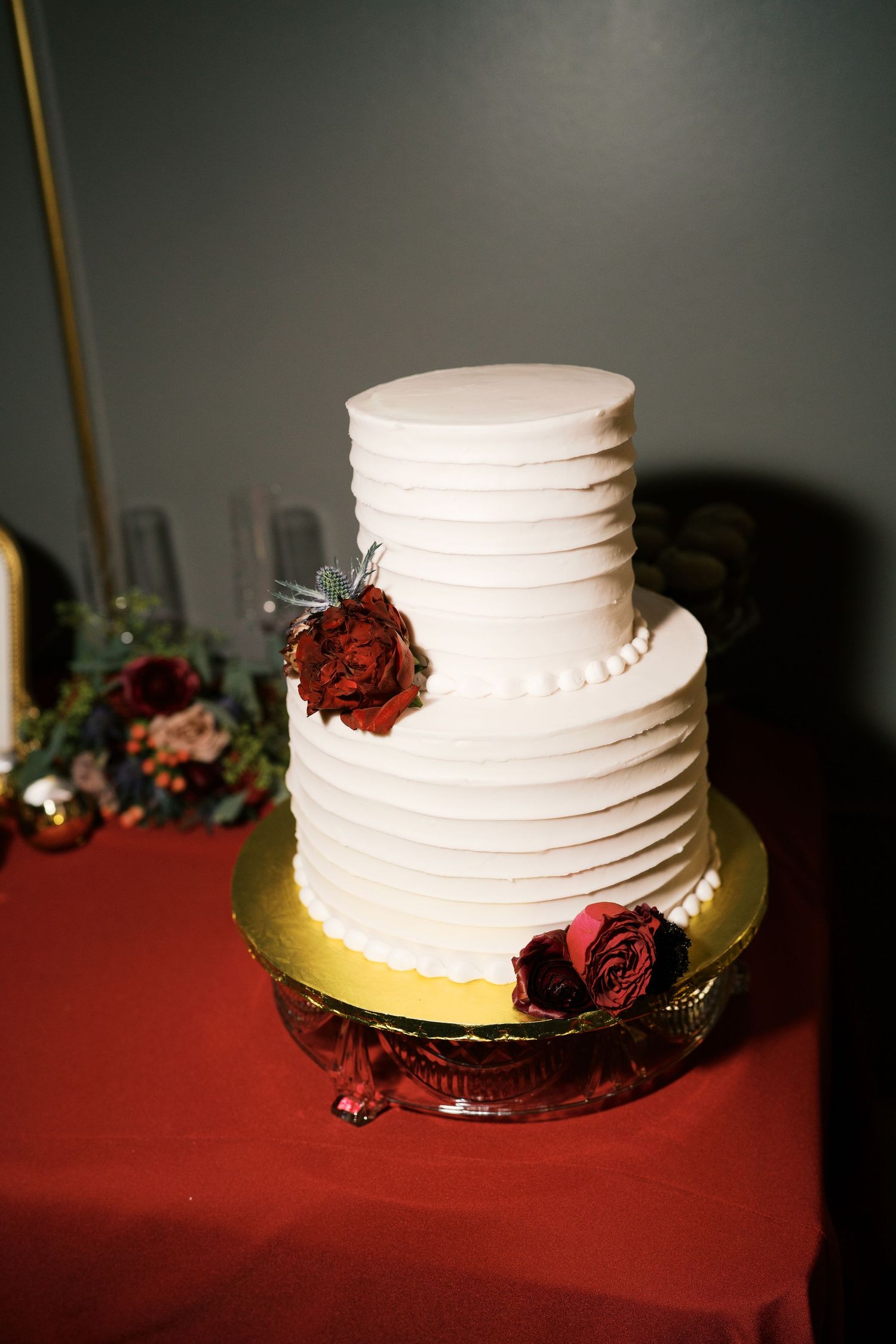 Elegant white wedding cake decorated with deep red roses on a burgundy tablecloth.