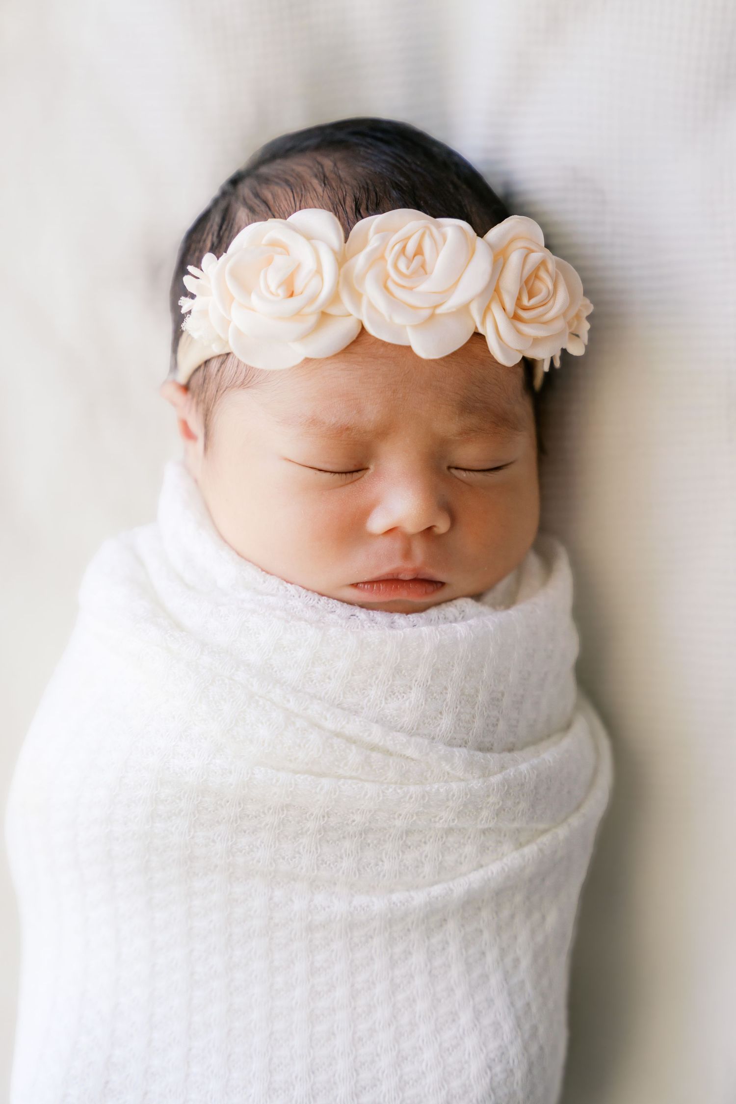 Newborn baby wrapped in white swaddle wearing a soft pink floral crown.