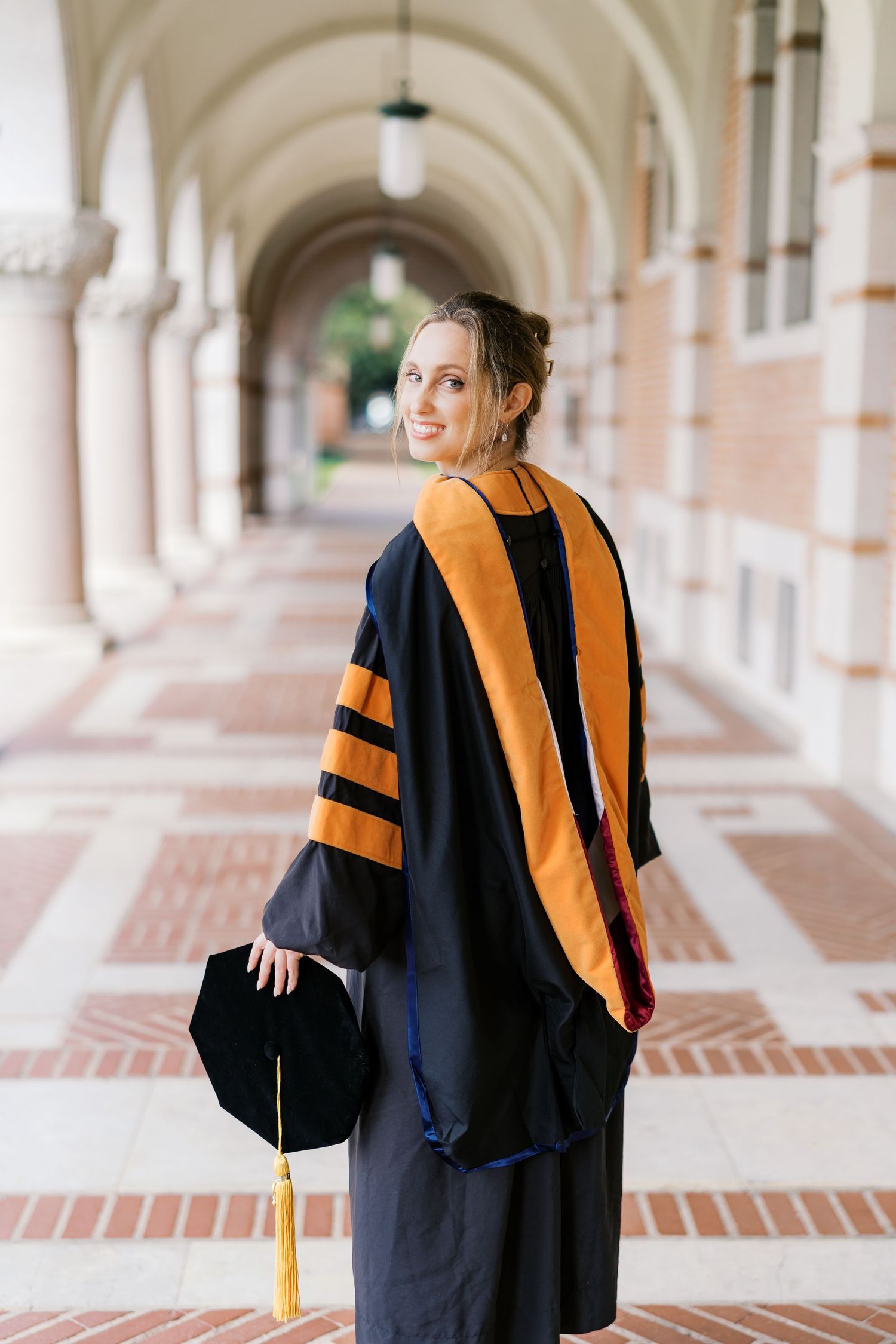Graduate in academic regalia with gold stole looks back while walking through university corridor.