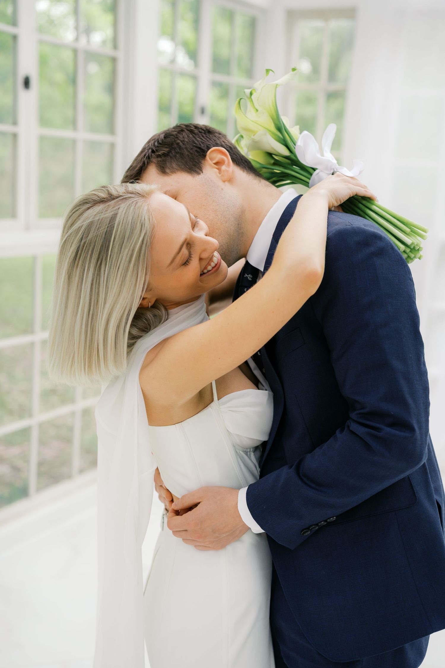 A bride and groom share a tender embrace, she holds white calla lilies, wearing a veil and white gown, he in a navy suit.