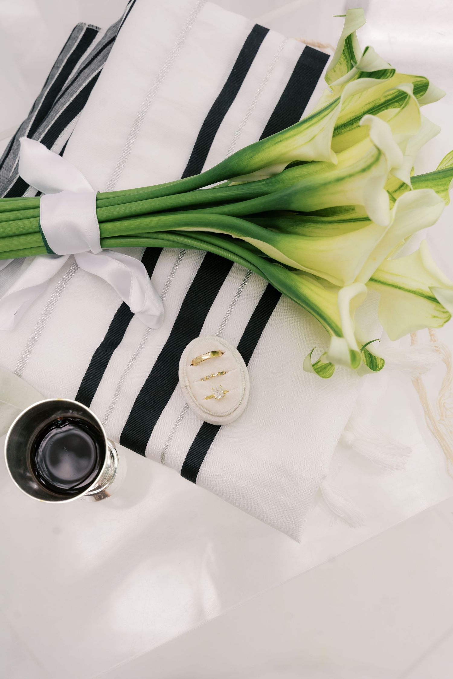 Elegant wedding flat lay with calla lily bouquet, gold rings, and striped tallit on white silk fabric.