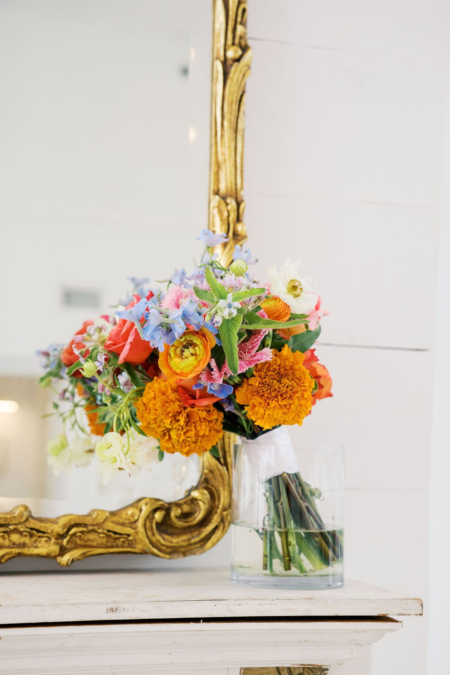 Colorful floral bouquet with orange marigolds, roses, and wildflowers in a glass vase on a white mantel with gold mirror.