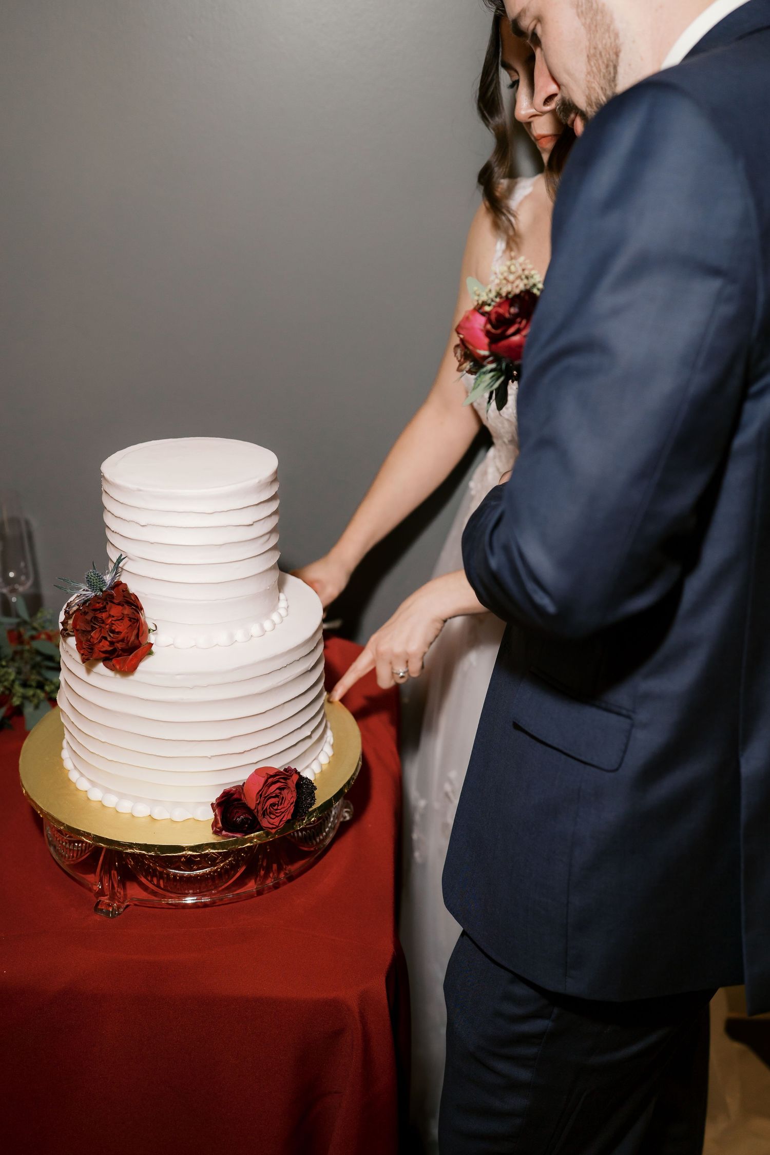 A white tiered wedding cake with red floral accents sits on a burgundy tablecloth.