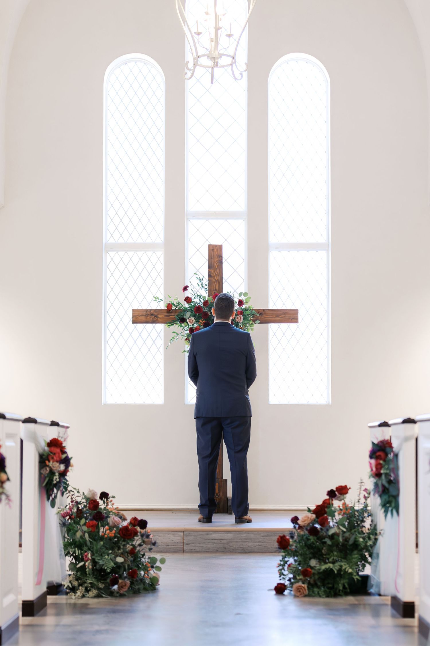 A couple shares their first look moment in a bright white church interior with red floral arrangements.