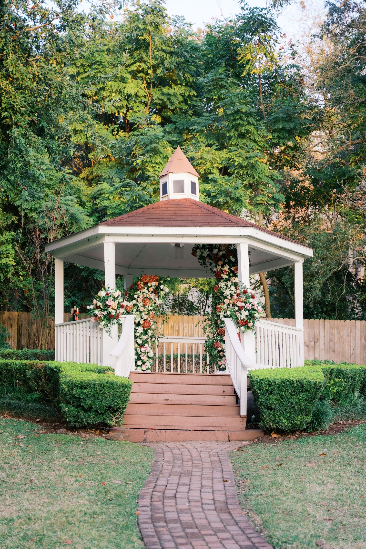 A charming white gazebo with brown roof connected by brick pathway in a garden setting.