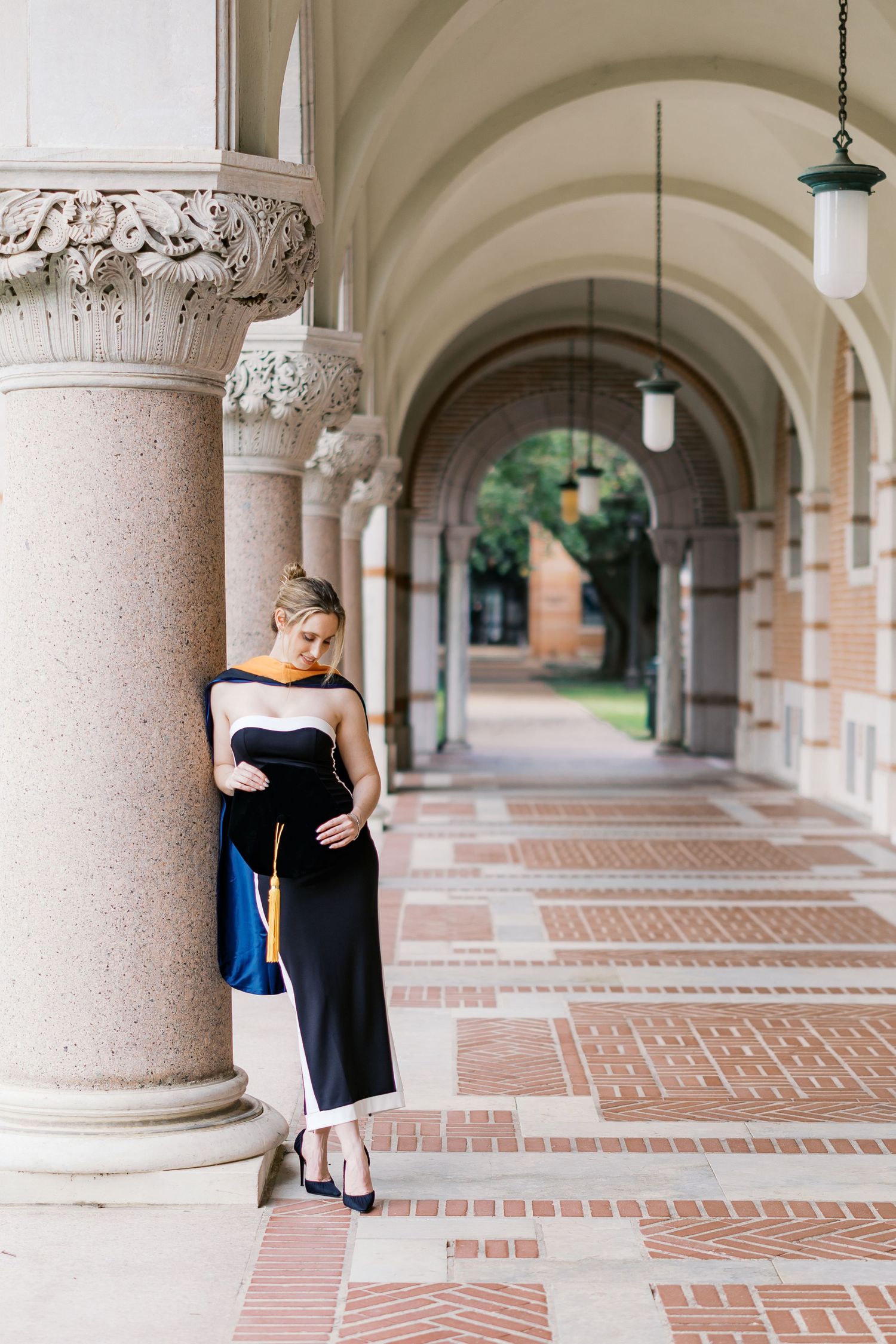 Graduate wearing black dress leans against decorative column in elegant archway.
