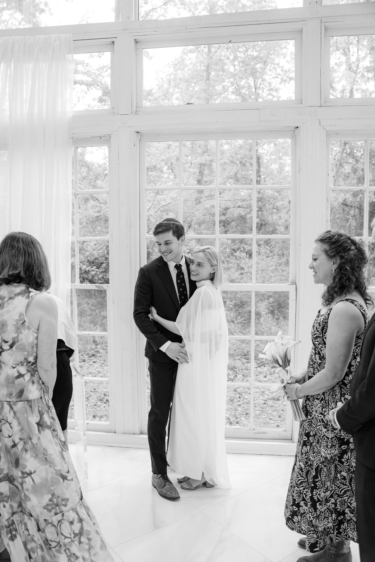 Black and white photo of a couple embracing at their wedding ceremony indoors, with guests looking on near large windows.