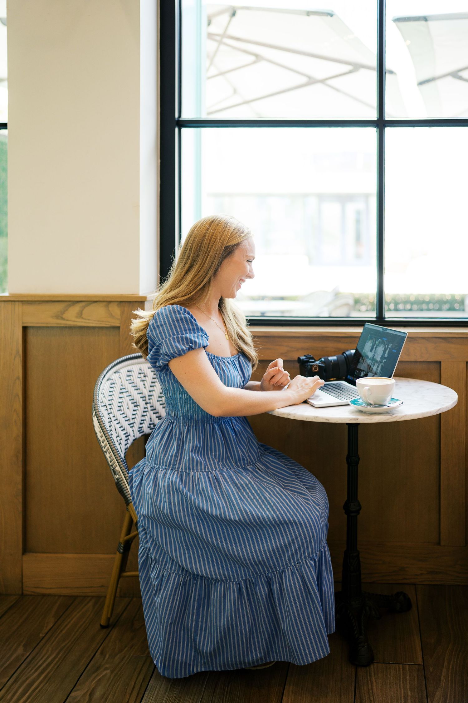 Woman in blue striped dress working at round table by window in cafe.