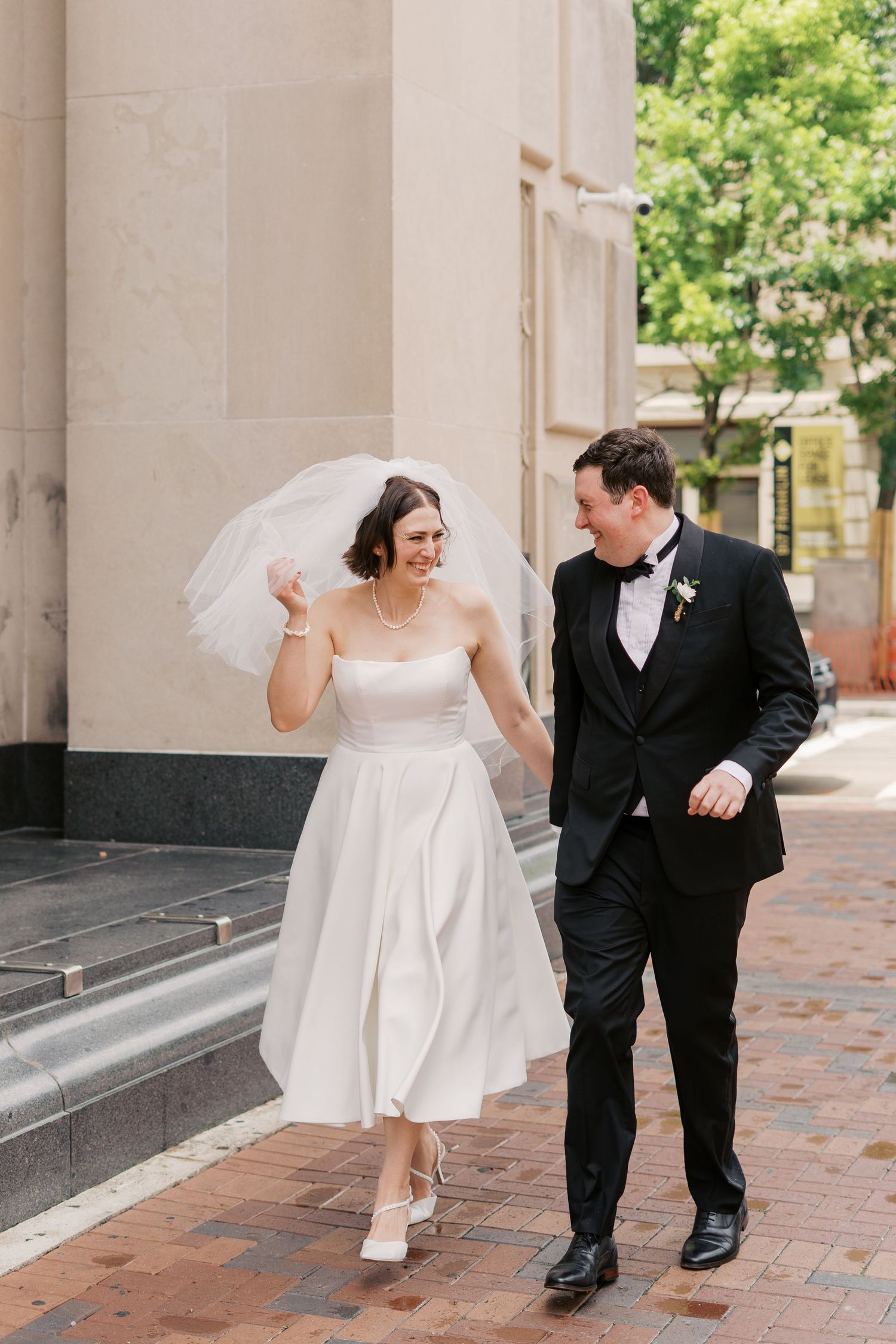 A bride and groom share playful moments walking together through a columned walkway.