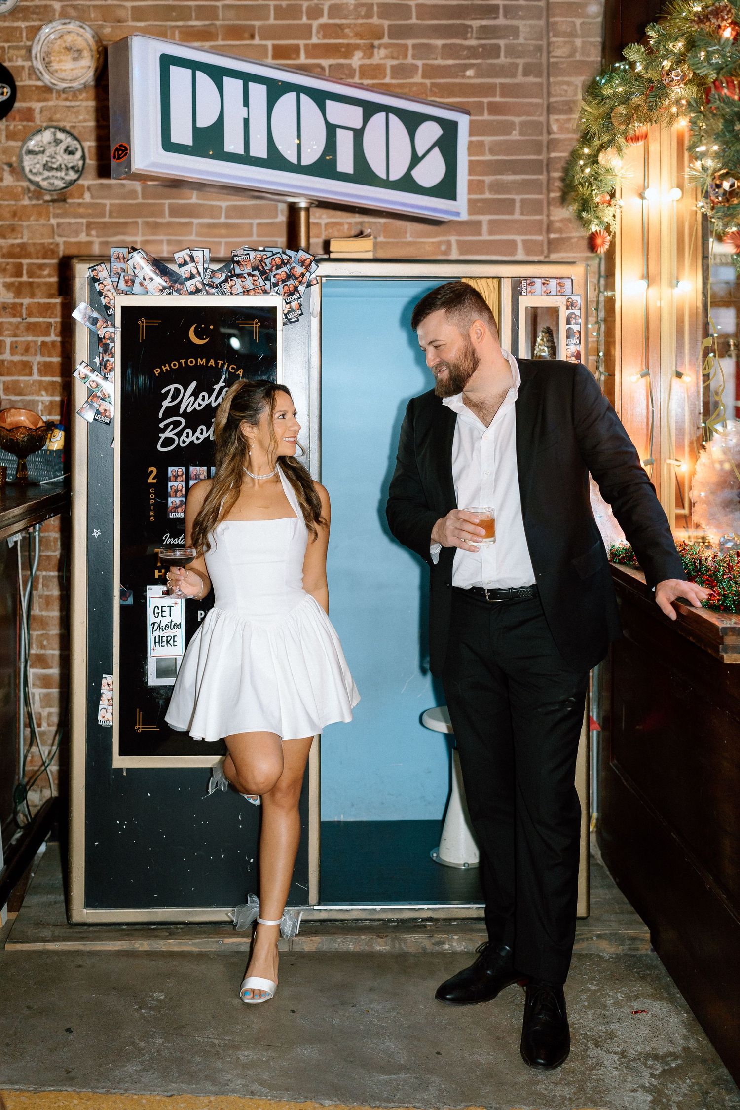 Couple in formal attire playfully interacting outside a brick storefront with neon signage at night.