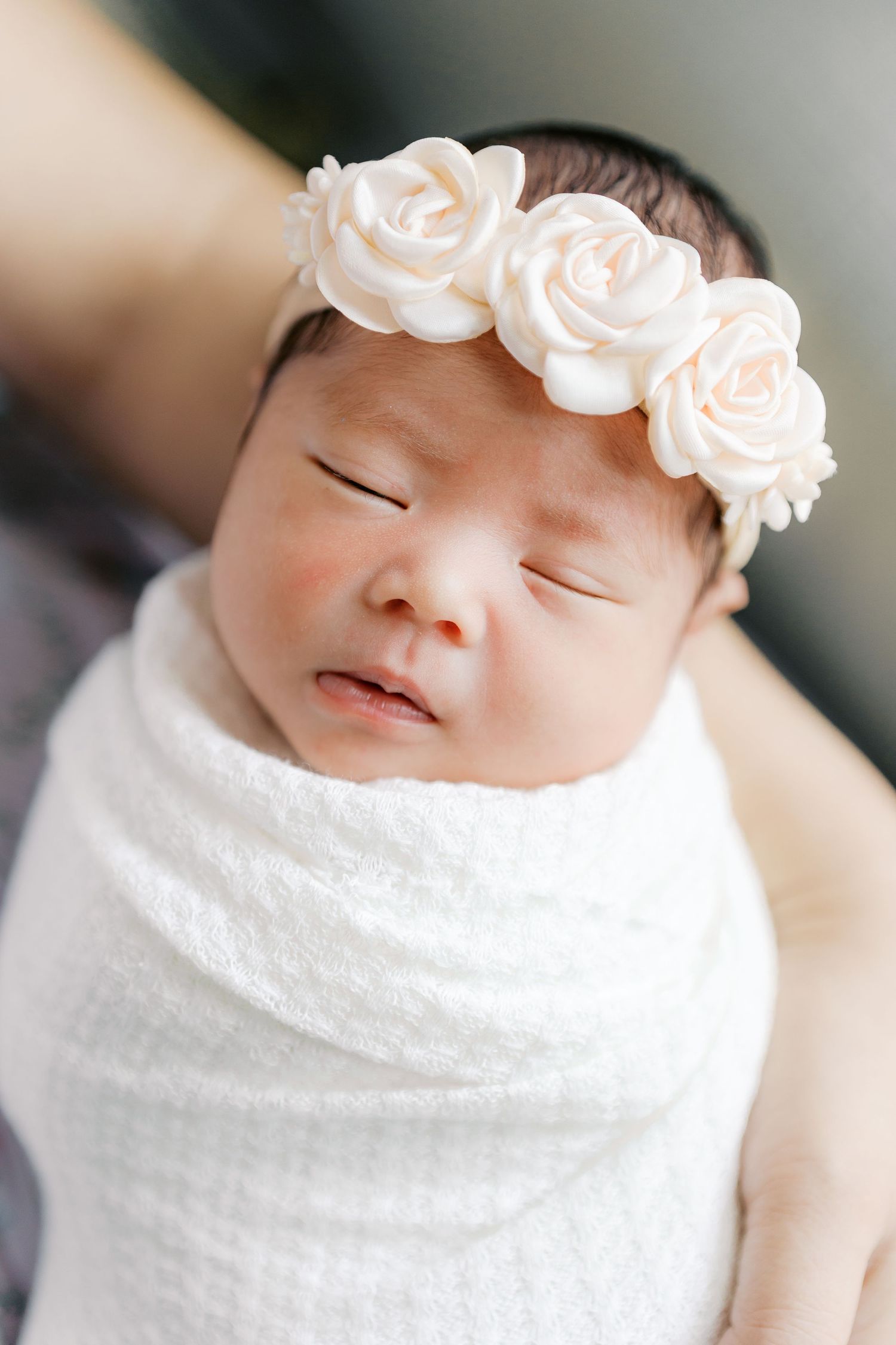 Close-up of peaceful sleeping newborn wearing white roses headband and swaddled in white fabric.