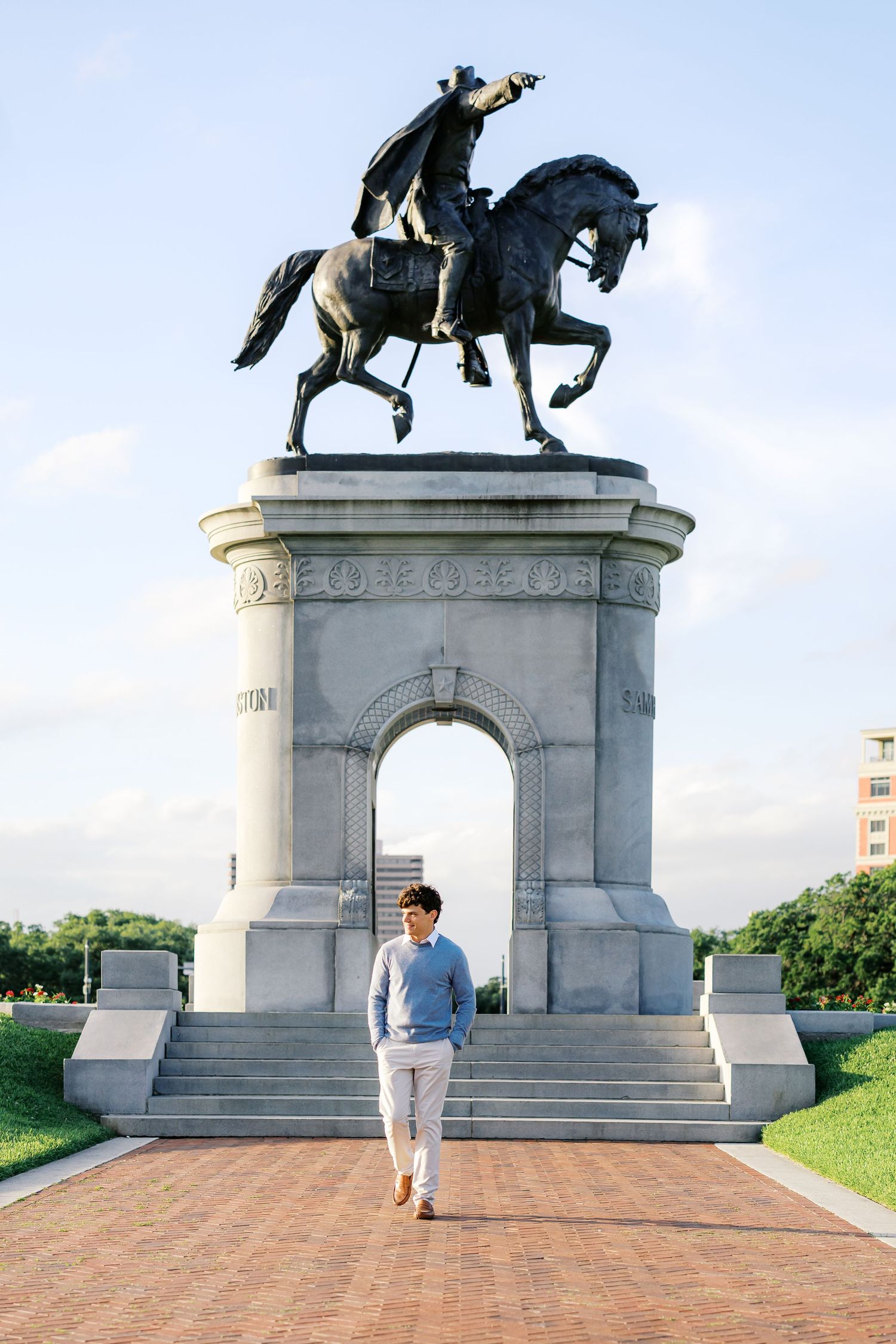 A young man walks beneath a grand equestrian statue monument on a brick pathway under a clear blue sky.