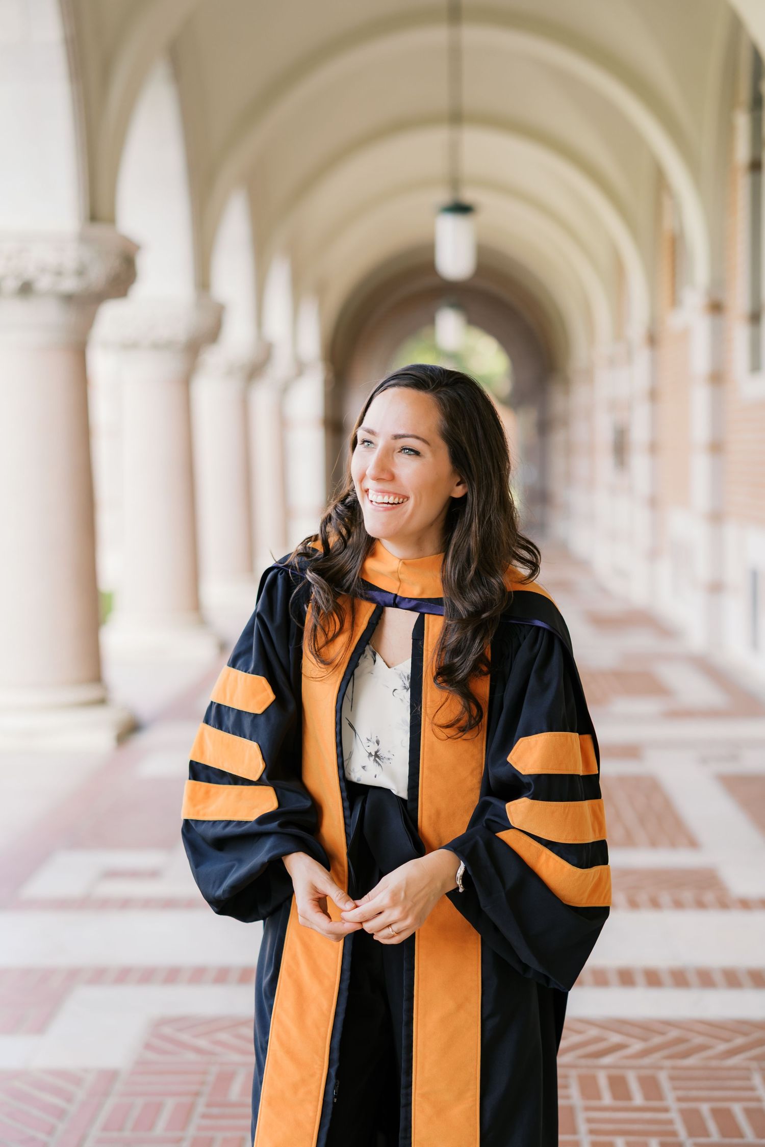 Graduate in black and orange academic regalia smiles while walking through an elegant columned hallway.