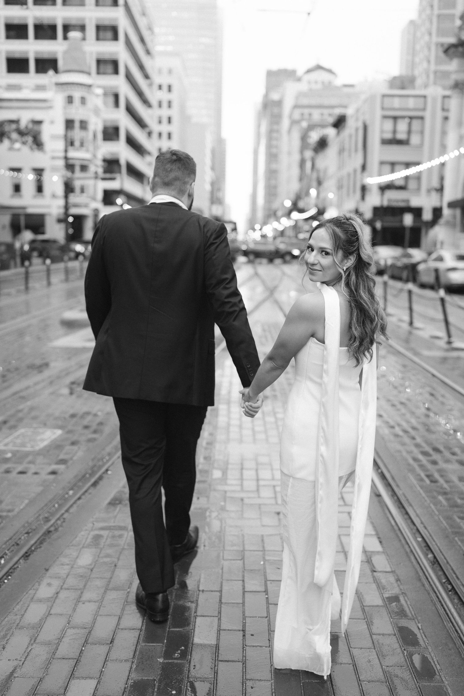 Elegant couple strolls down wet cobblestone street with trolley tracks.