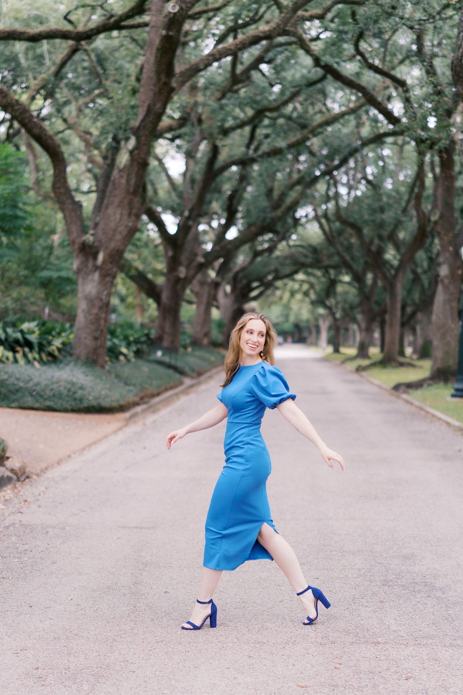 A figure in a bright blue dress and blue heels dances playfully on a tree-lined path.