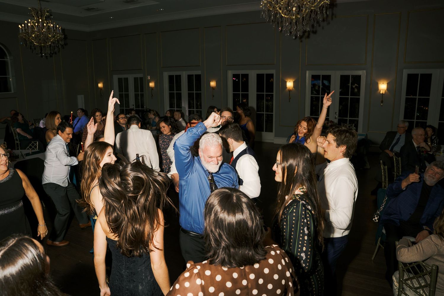 Wedding guests dancing with raised hands under elegant chandeliers at an indoor reception.