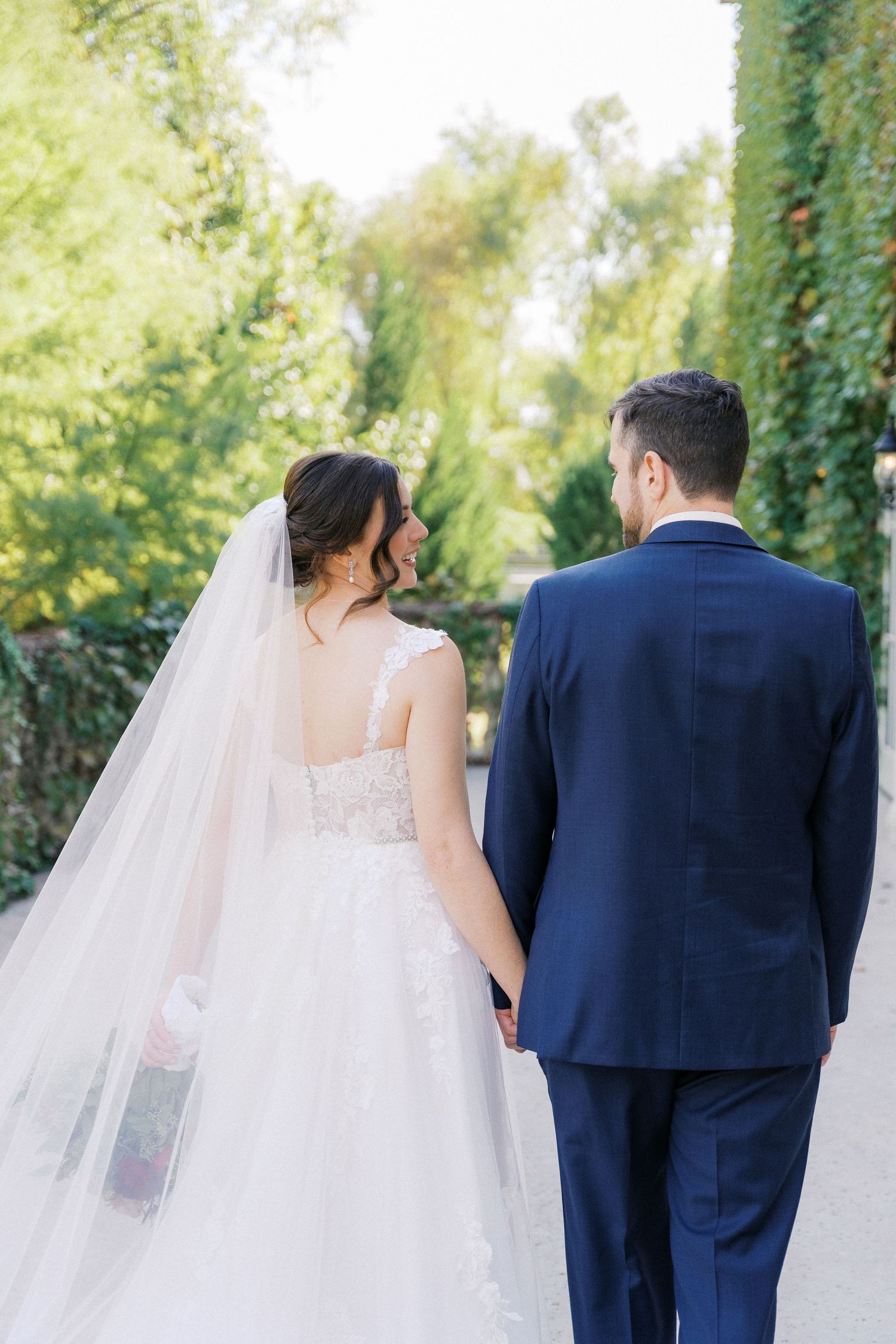 Newlyweds walking together on a stone path with trailing wedding veil and green foliage backdrop.