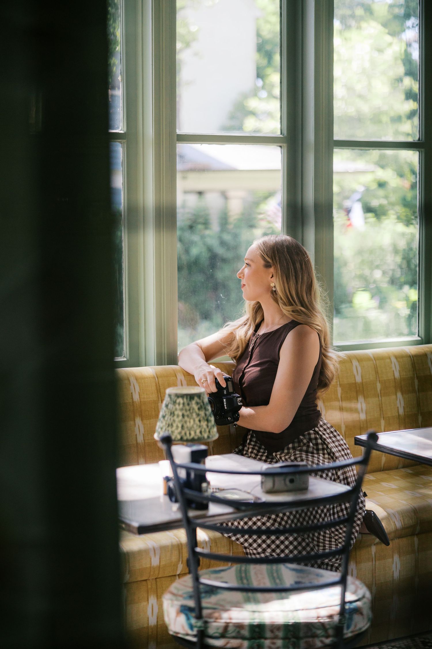 Blonde woman sitting by a sunlit cafe window, holding a camera, wearing a dark top and patterned skirt.