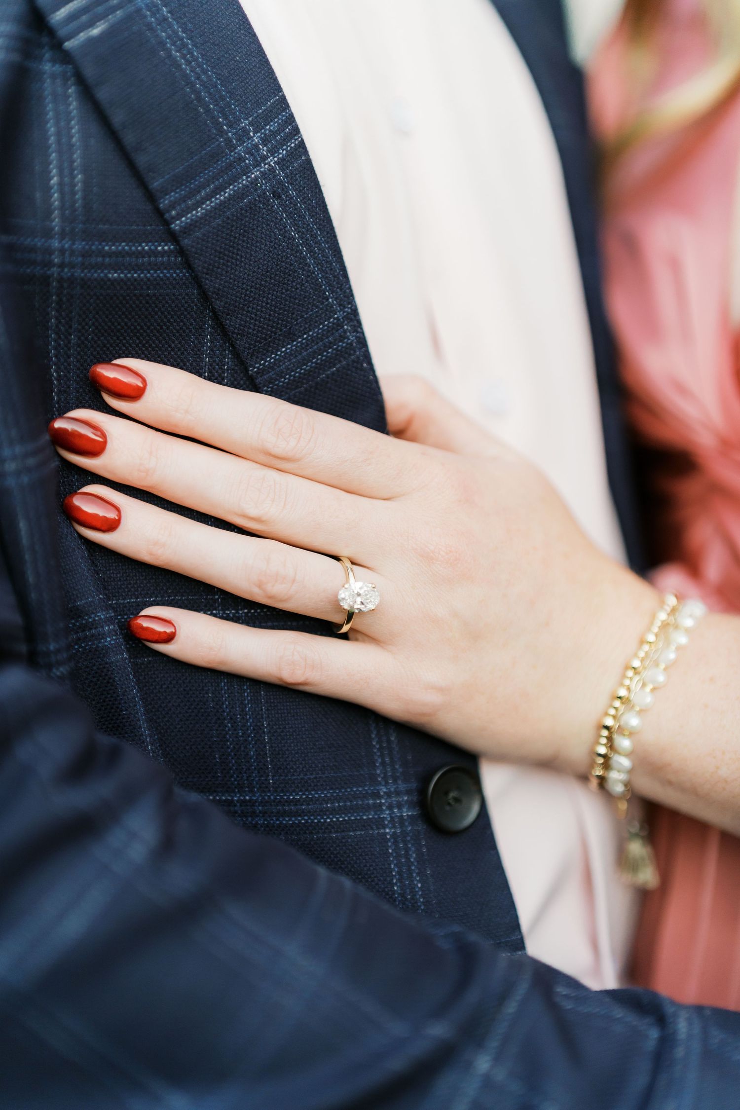 Close-up of engagement ring and red manicured nails against navy suit jacket.