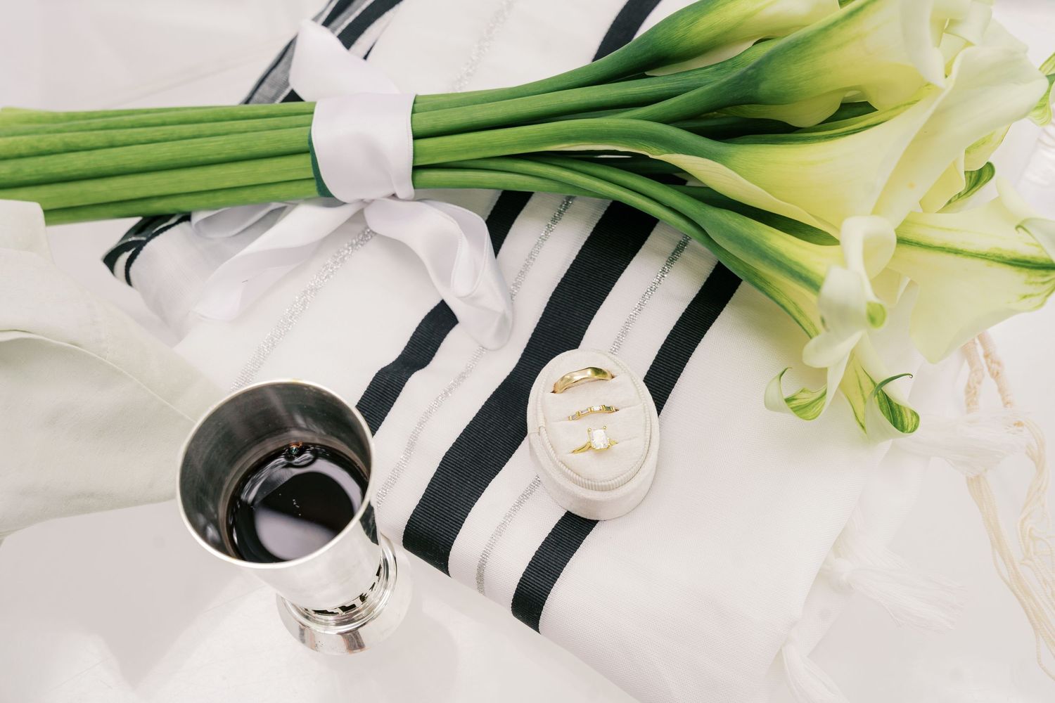 Jewish wedding essentials: tallit prayer shawl, silver kiddush cup with wine, gold rings, and white calla lily bouquet.